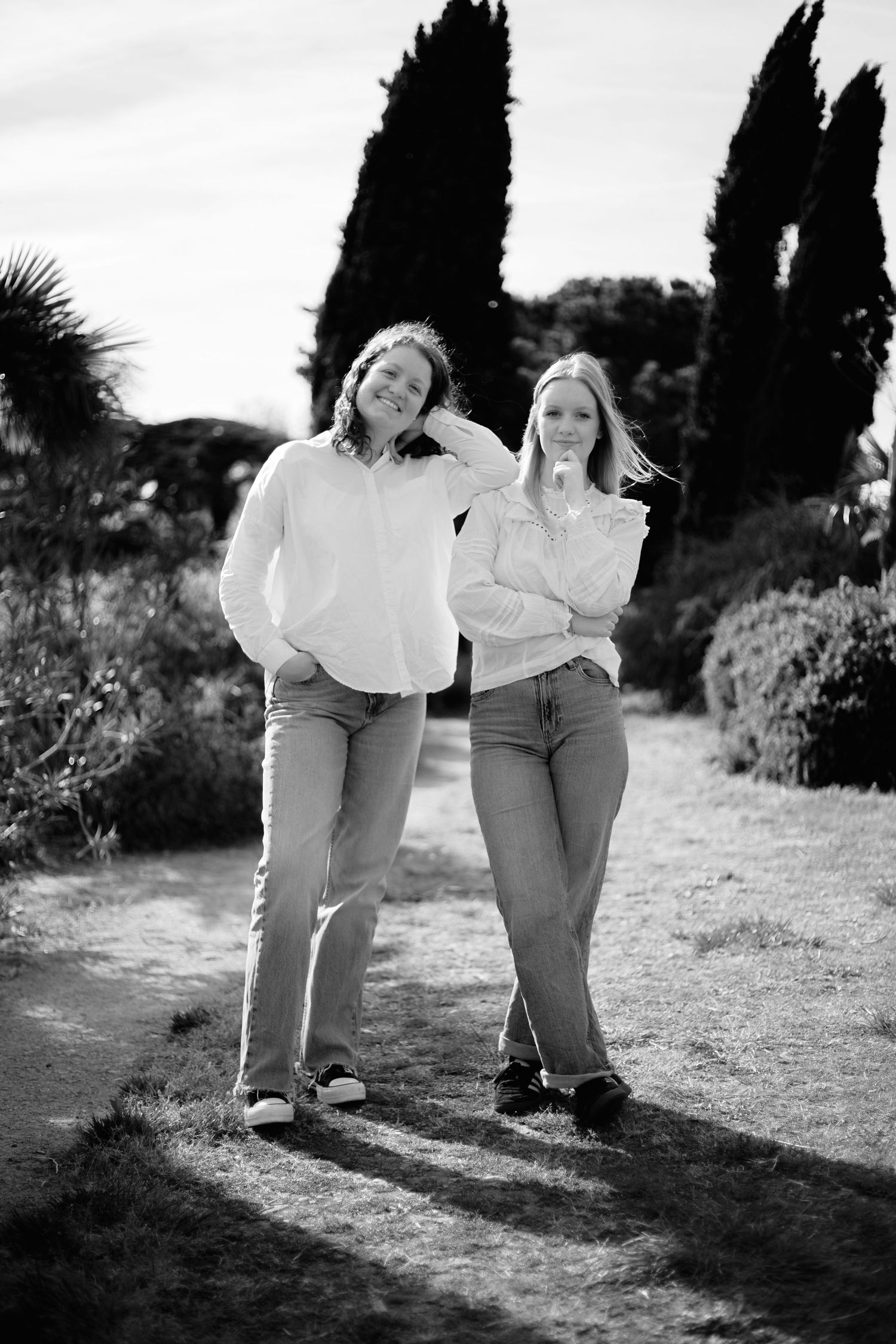 Black and white photo of two women in white sweaters and jeans posing outdoors among trees and shrubs.