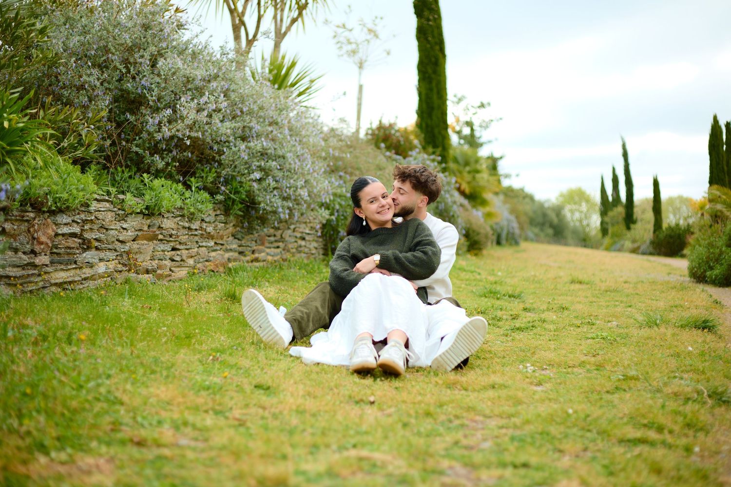 Couple in wedding attire sitting together on green grass surrounded by trees and a stone wall.