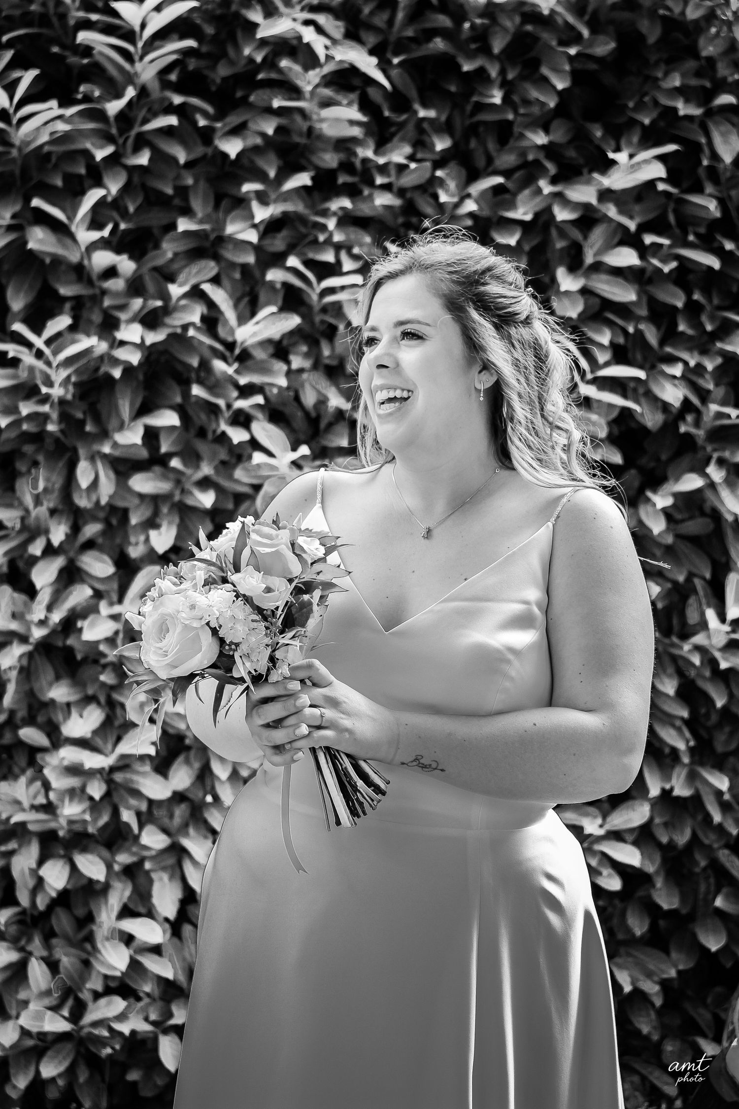 A black and white portrait of a bride holding a bouquet against a leafy backdrop.