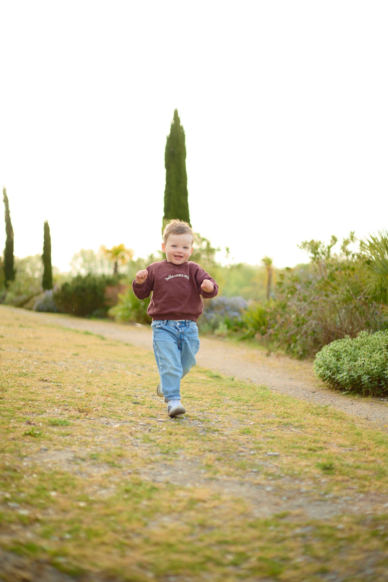 A happy young boy running along a garden path wearing jeans and a maroon sweatshirt on a bright day.
