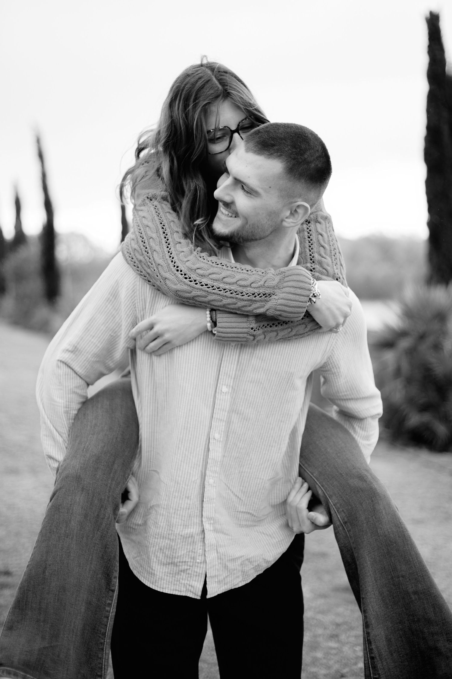 Black and white photo of a smiling man giving a woman a piggyback ride outdoors near trees.