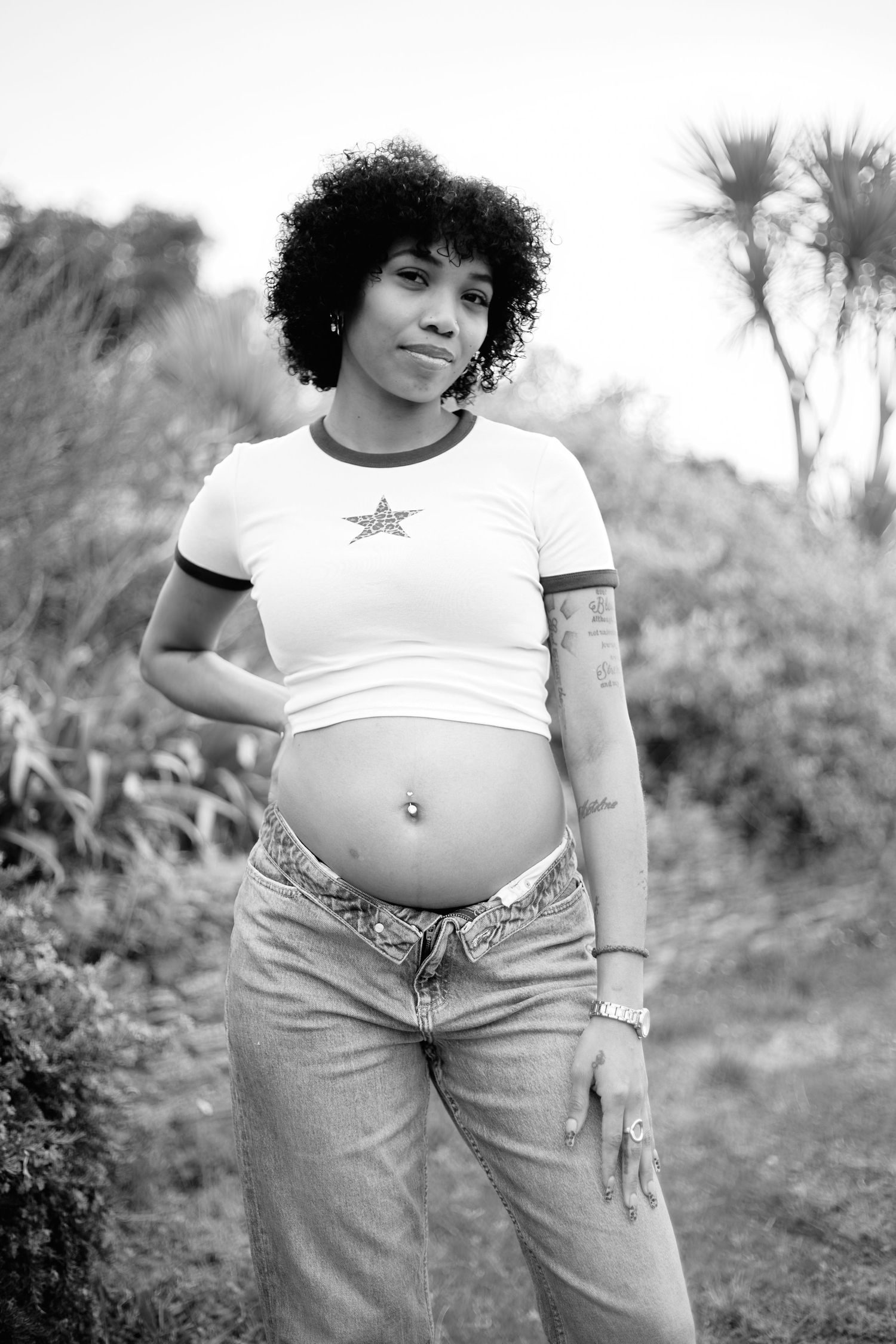 Pregnant woman with curly hair poses outdoors in crop top and jeans, showing baby bump in black and white photo.