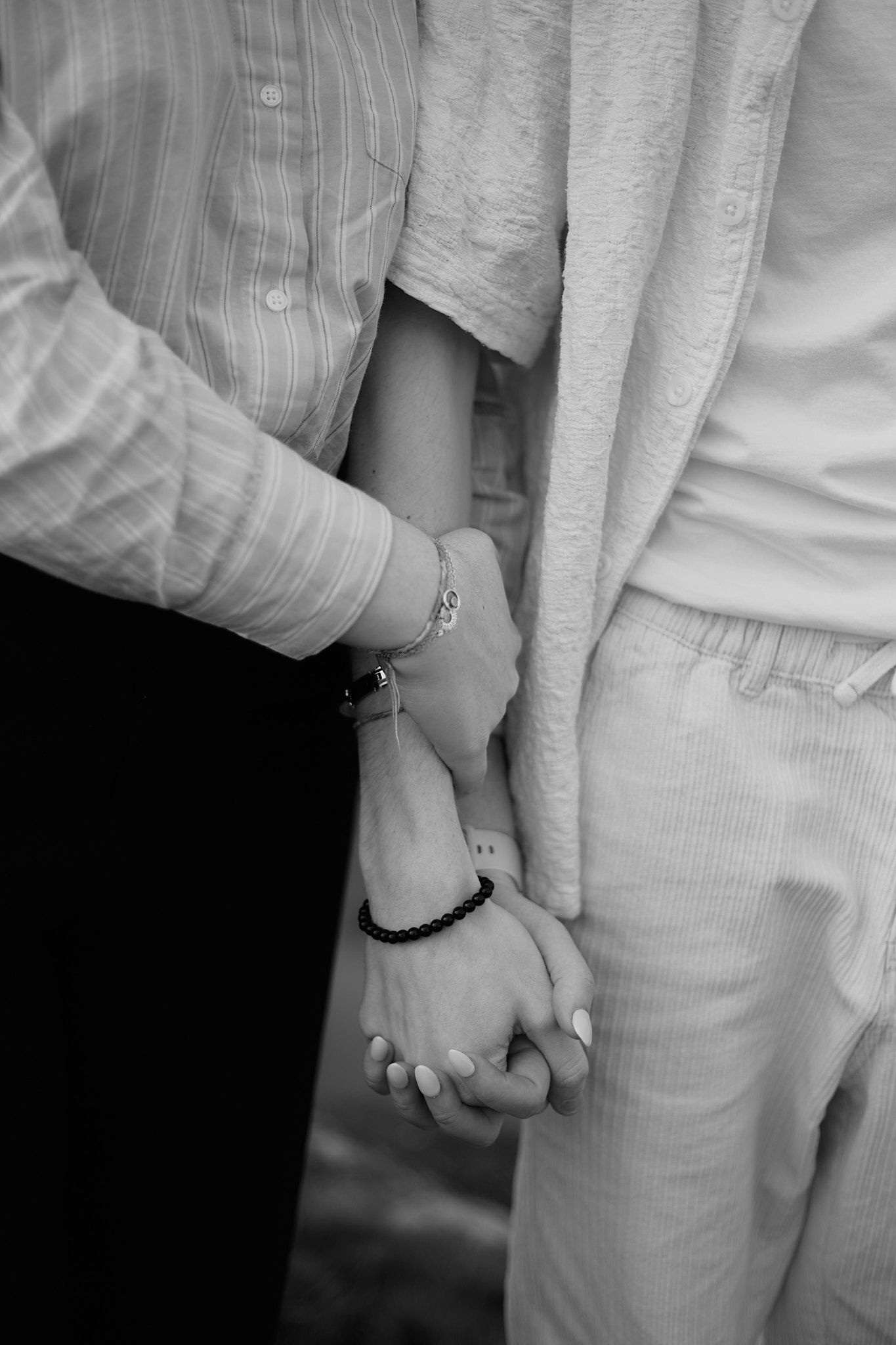 Black and white close-up of two people holding hands intimately with bracelets and rings visible.