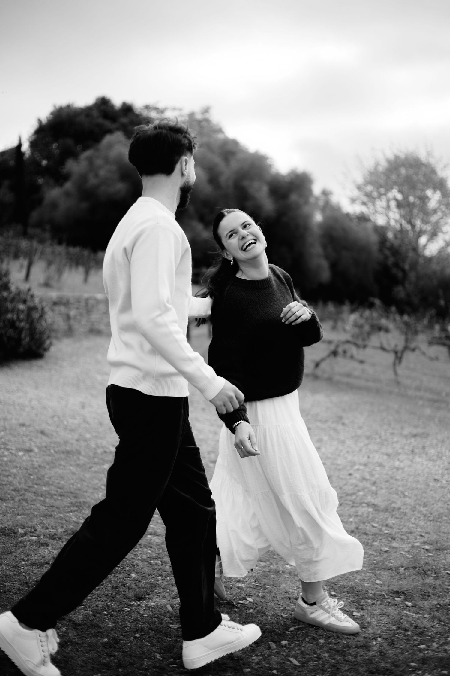 Black and white photo of a happy couple holding hands and laughing while walking outdoors among trees.
