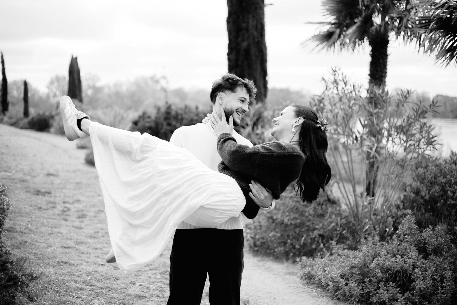 Black and white photo of a groom lifting his bride in a flowing white dress outdoors near palm trees.