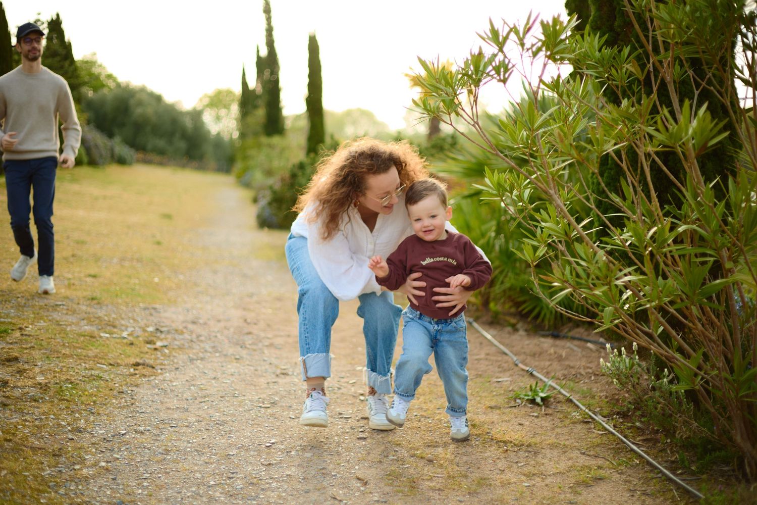 A mother helps her toddler walk on a garden path while a man strolls behind them outdoors.