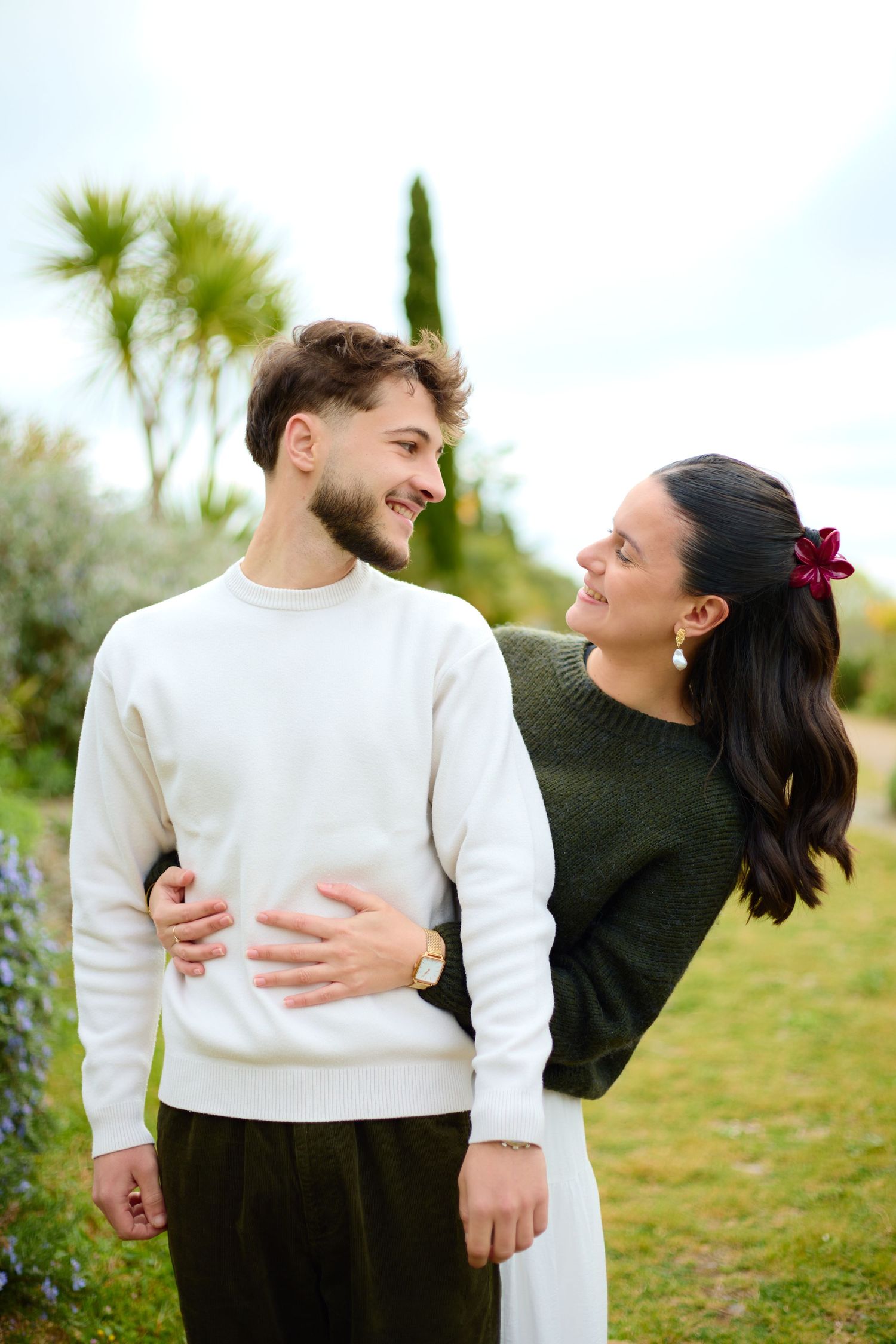 Happy young couple embracing outdoors in a lush garden, smiling at each other on a cloudy day.