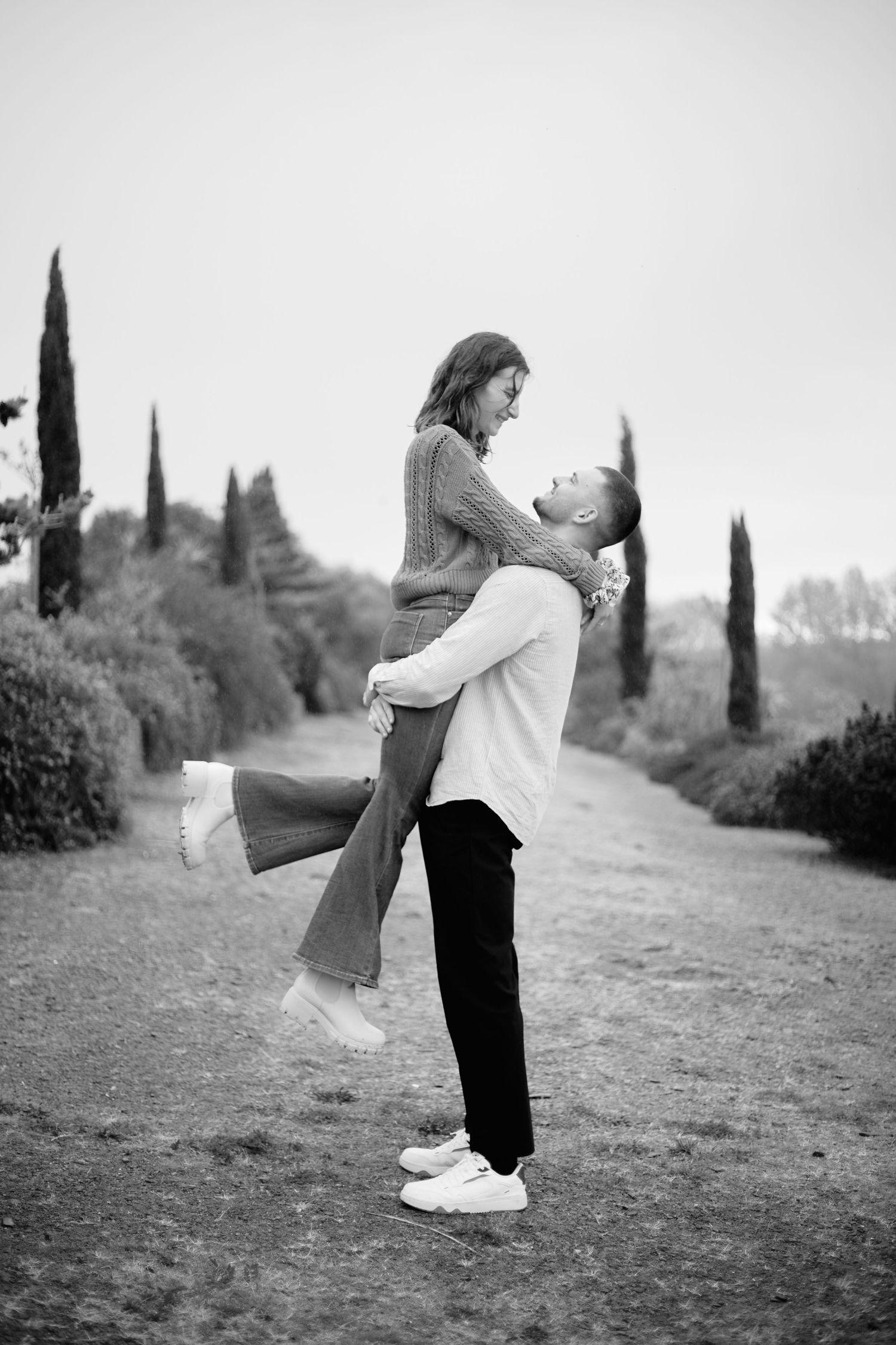 Black and white photo of a man lifting a woman on a tree-lined path, sharing a romantic moment outdoors.
