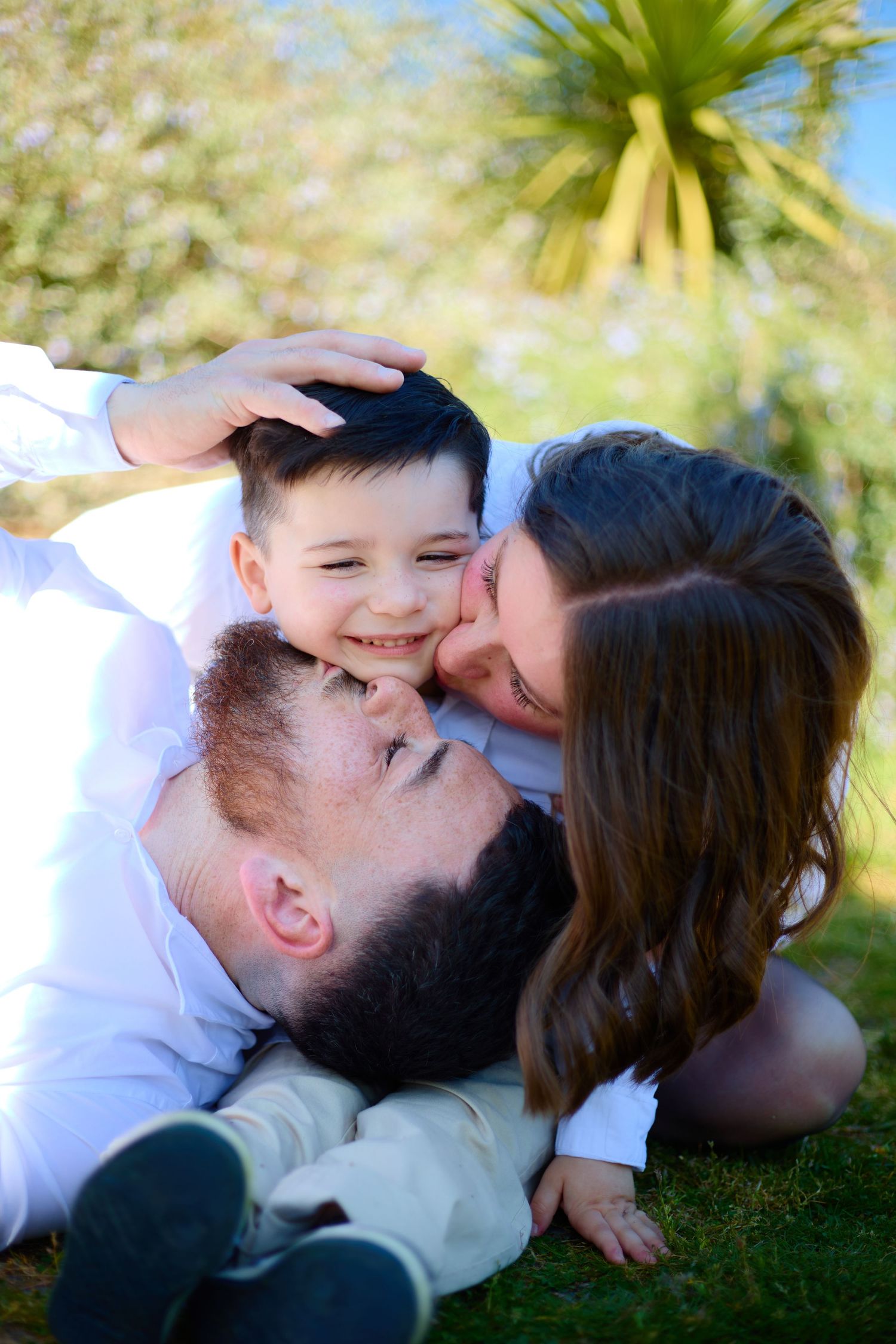 Happy family moment with parents kissing young smiling boy on cheeks while relaxing on green grass outdoors.