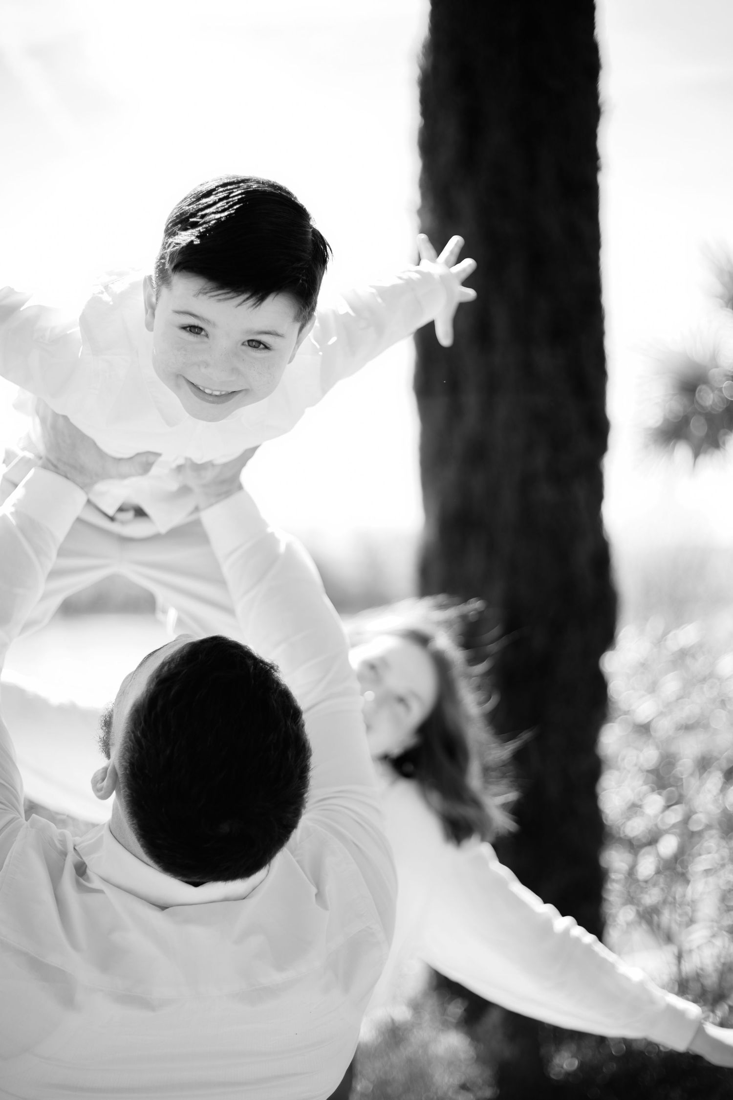Black and white photo of a joyful child being lifted overhead by parents outdoors near trees.