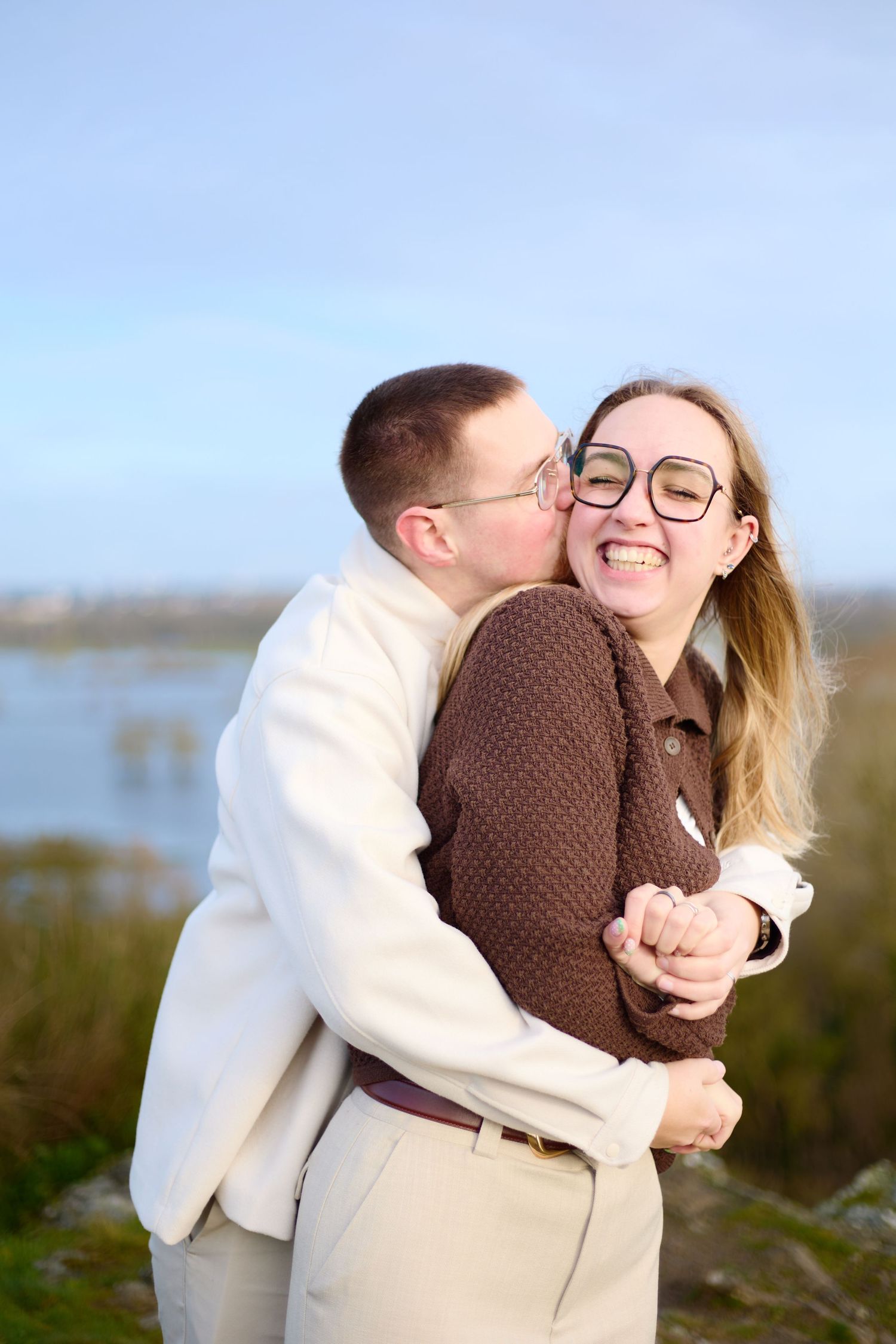 Couple embracing outdoors against blue sky with one wearing brown sweater and other in cream jacket.
