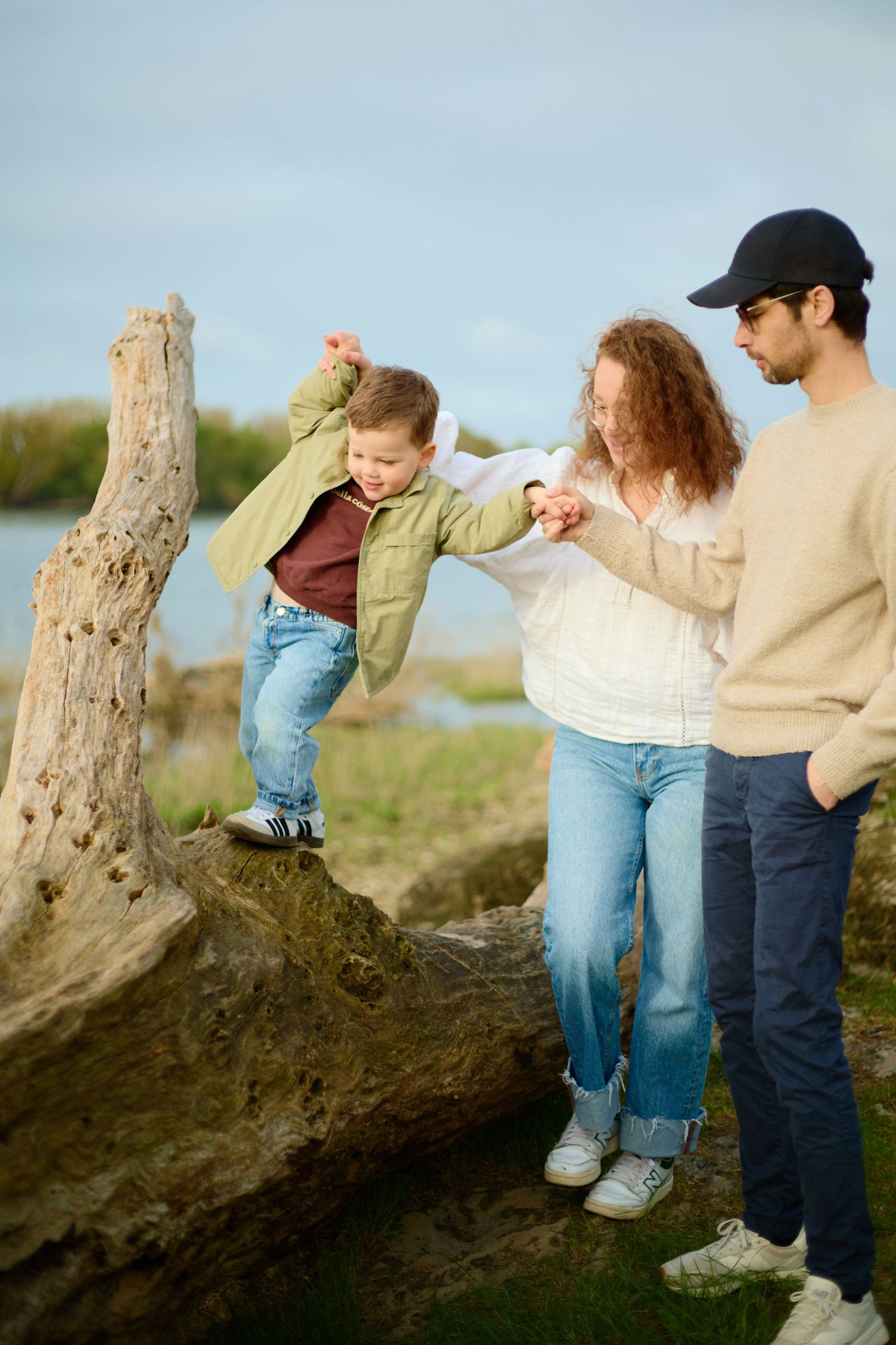 Family helping young boy climb driftwood log near water, parents assisting child outdoors on cloudy day.
