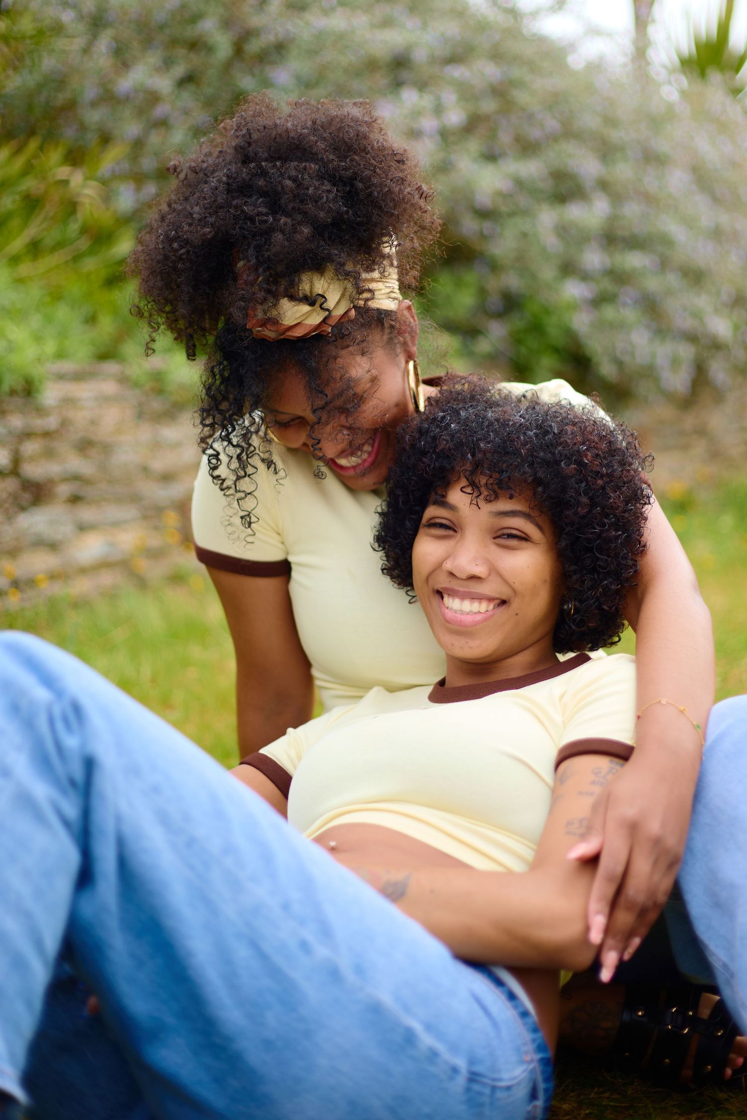 Two young Black women with curly afros sit together outdoors, smiling and embracing in a garden setting.