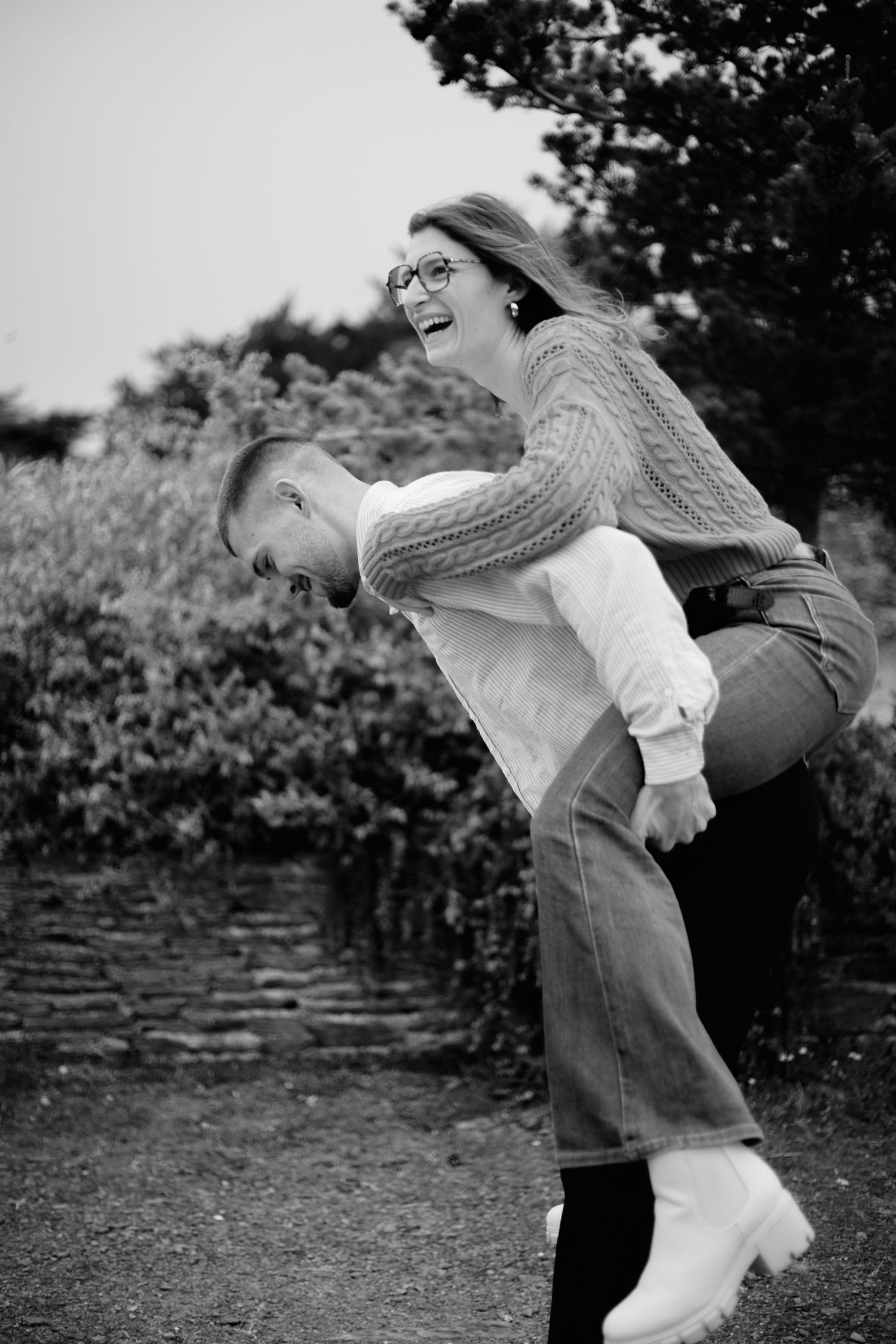 Black and white photo of a laughing couple, man giving woman a piggyback ride outdoors near lush greenery.