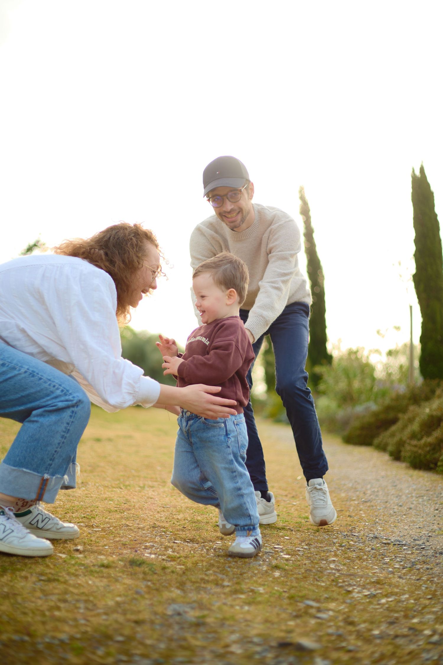 A young family with a toddler taking first steps outdoors in a sunny park, parents smiling and encouraging the child.