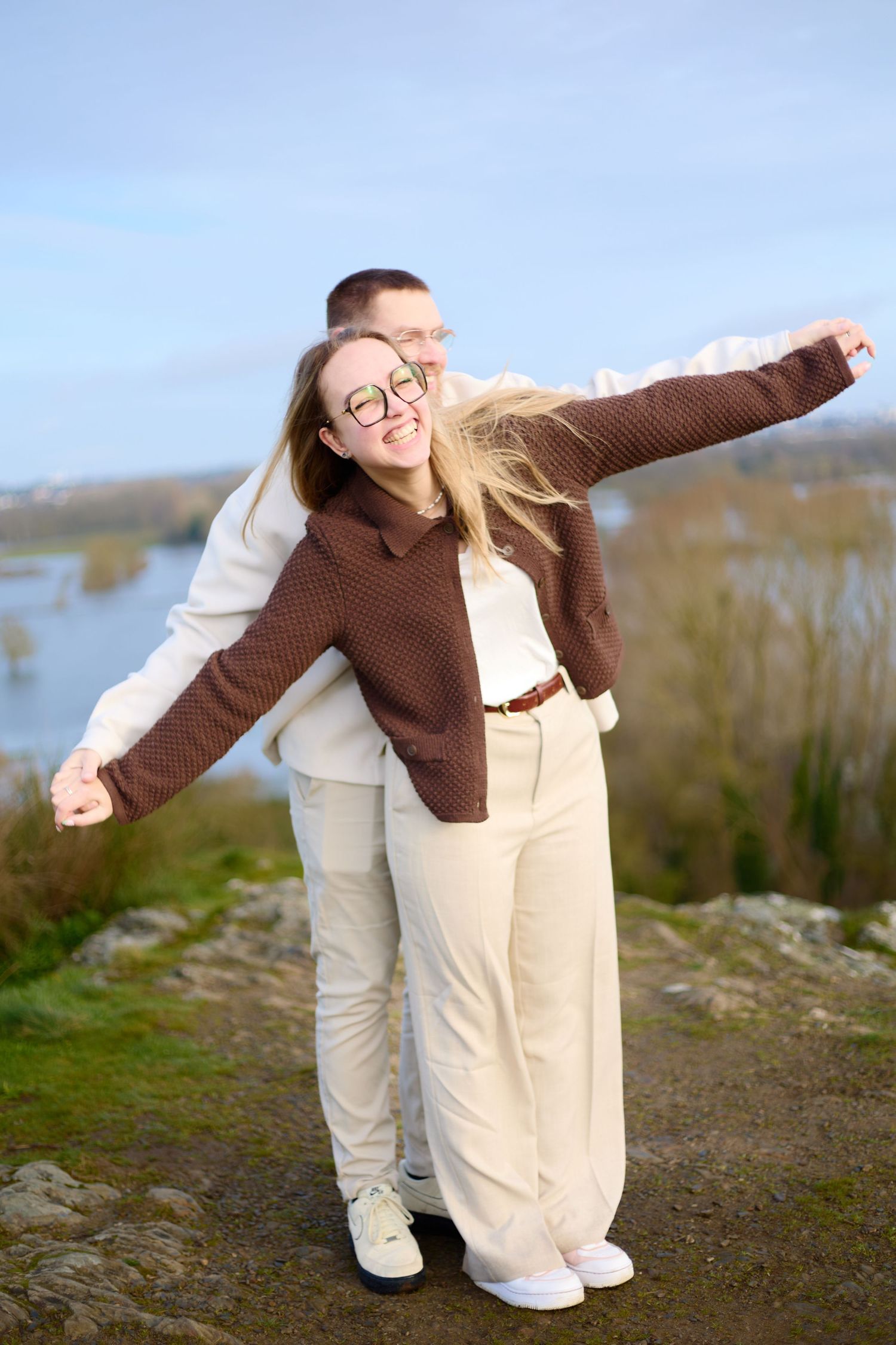 Playful couple in coordinated cream and brown outfits posing outdoors with arms spread wide.