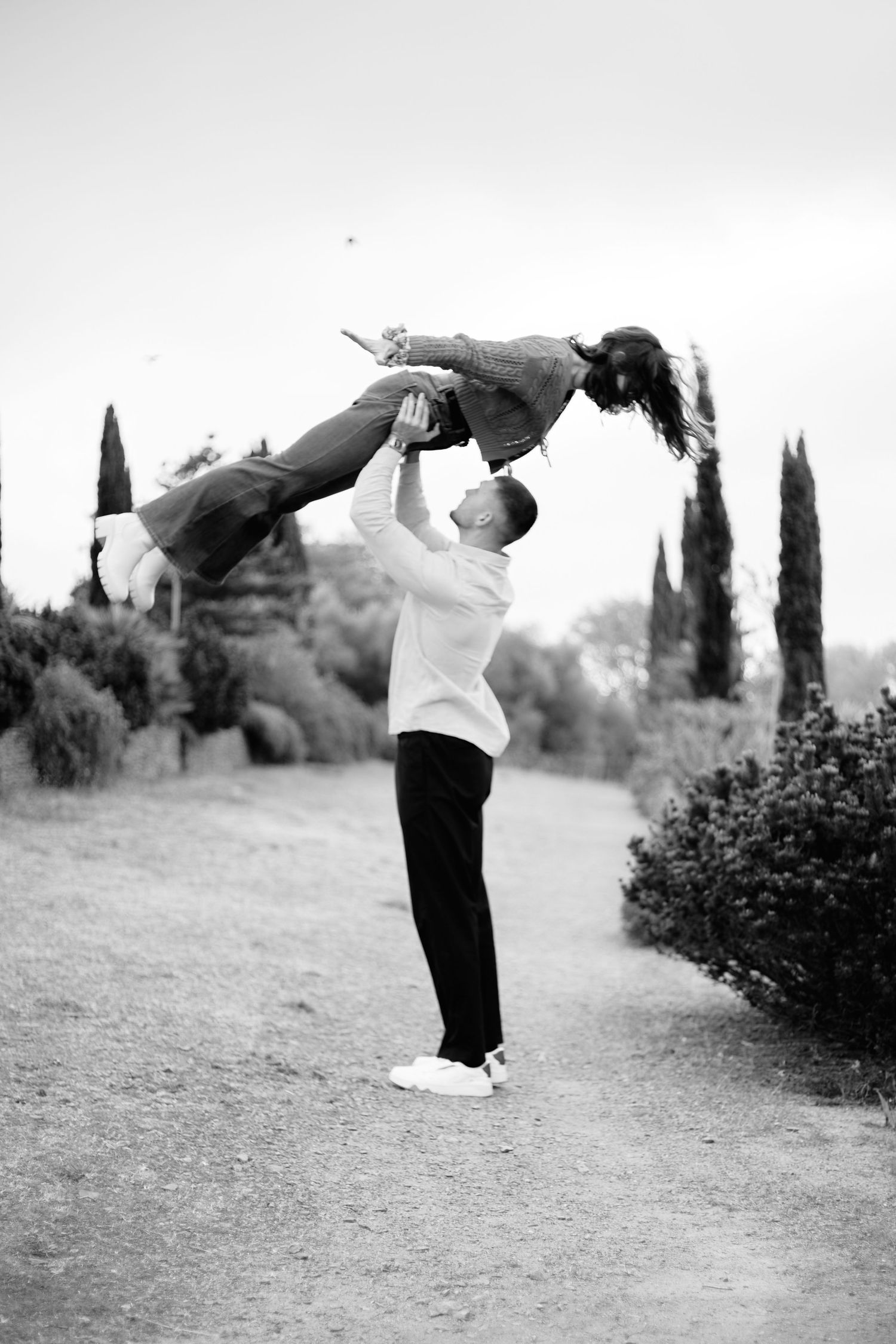 Black and white photo of a man lifting a child high in the air on a tree-lined path outdoors.