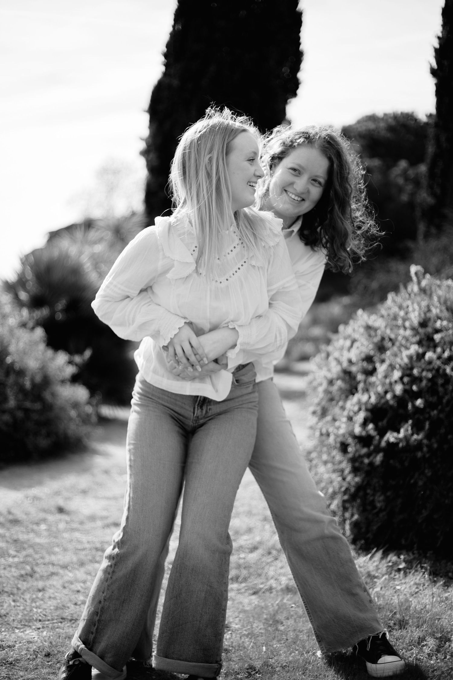 Two young women in white blouses and jeans embrace and laugh together in a black and white outdoor garden photo.