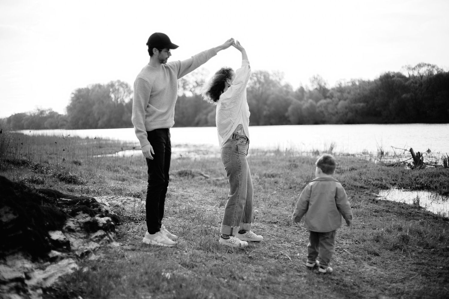 Black and white photo of a couple dancing outdoors near a lake while a toddler watches, capturing a joyful family moment.