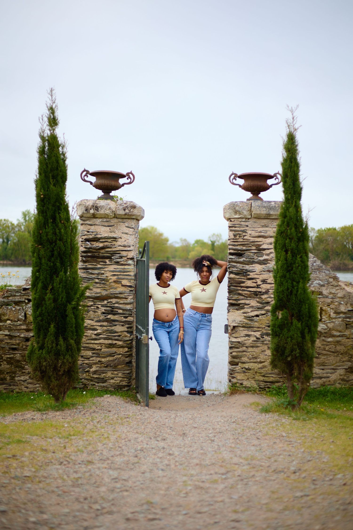 Two women in matching outfits pose at a stone gate with cypress trees and decorative urns on a cobblestone path.