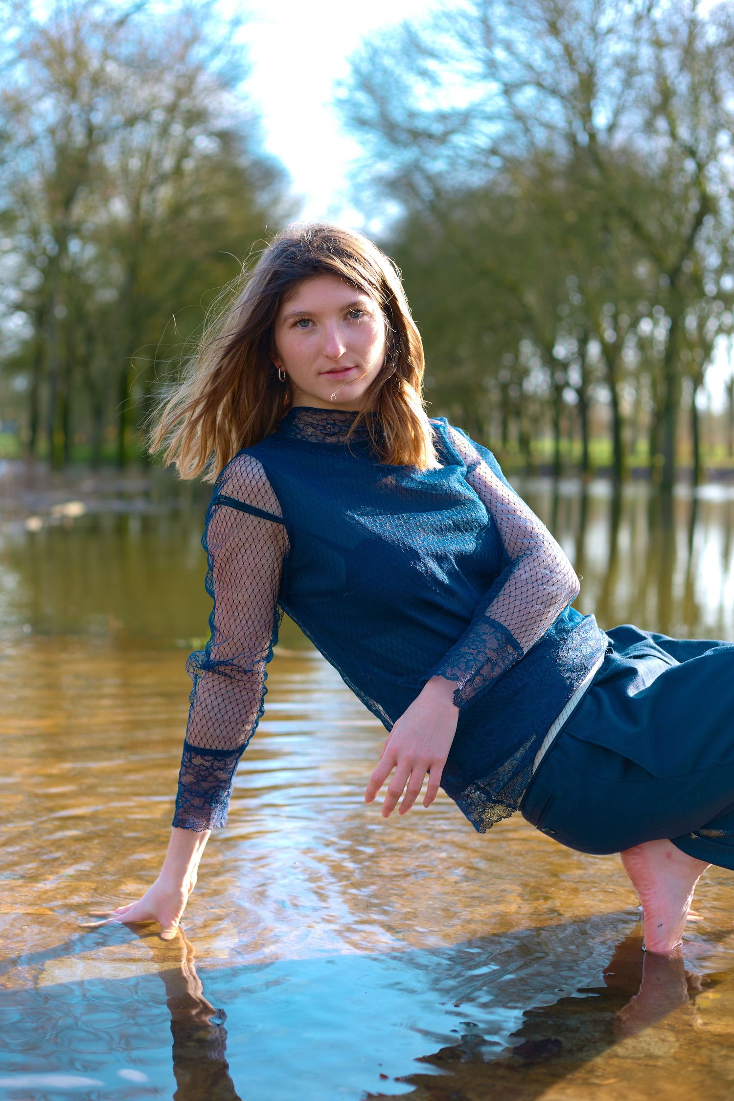 Person in blue dress poses artistically by water with trees reflected in background.
