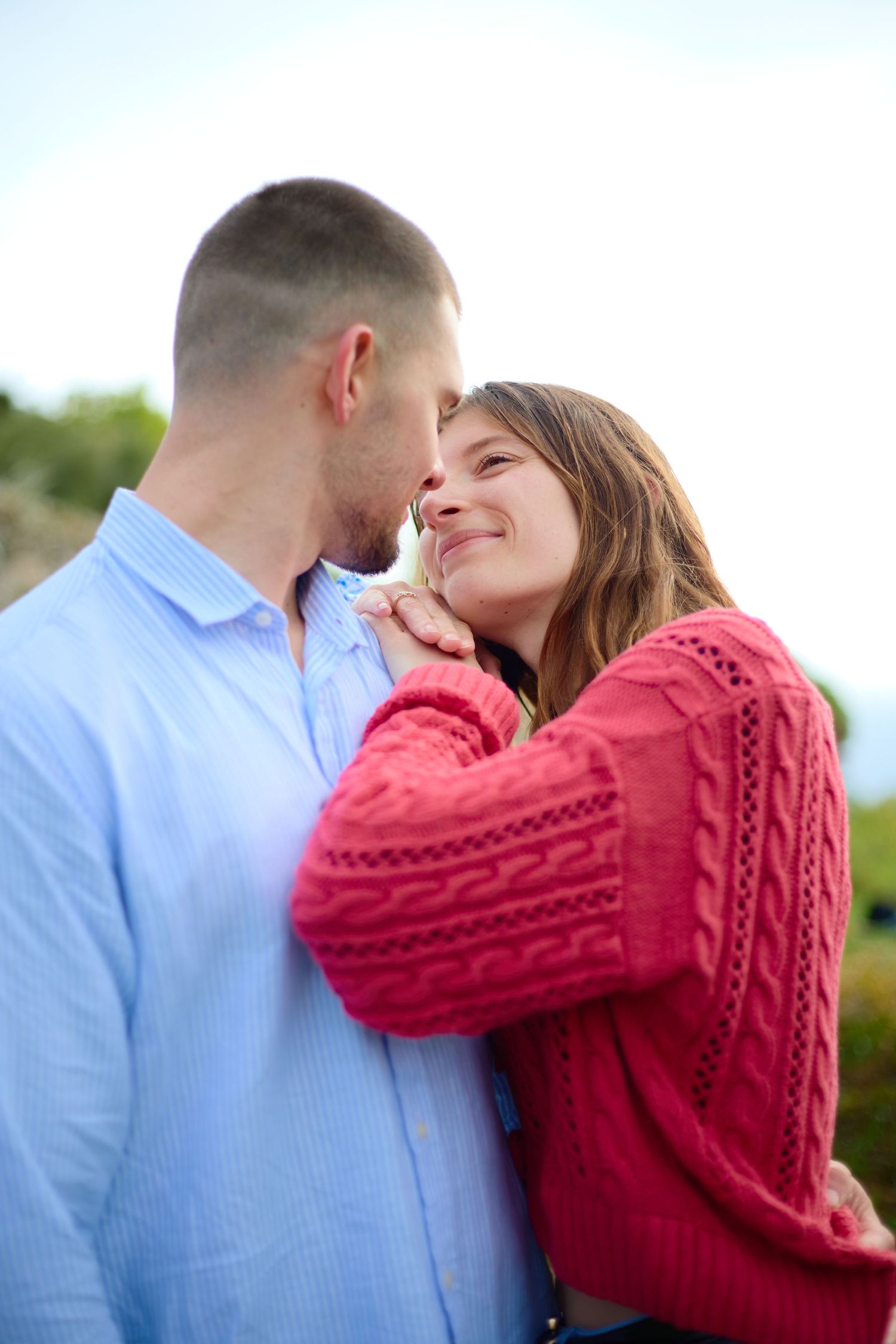 Romantic couple in casual wear sharing a tender moment outdoors, woman in red knit sweater smiling up at man in blue shirt.