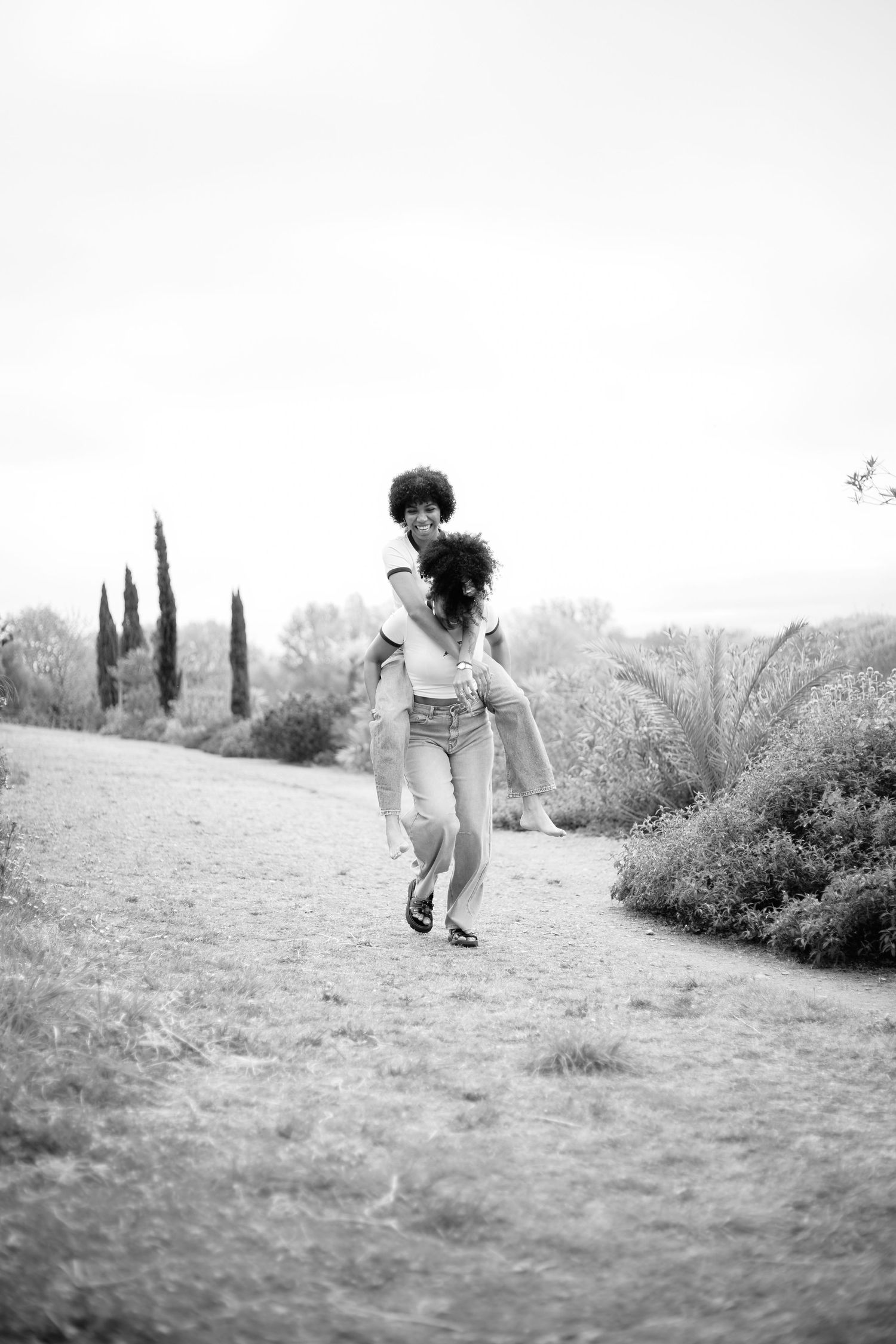 Black and white photo of a couple playing piggyback on a scenic outdoor path surrounded by trees and shrubs.