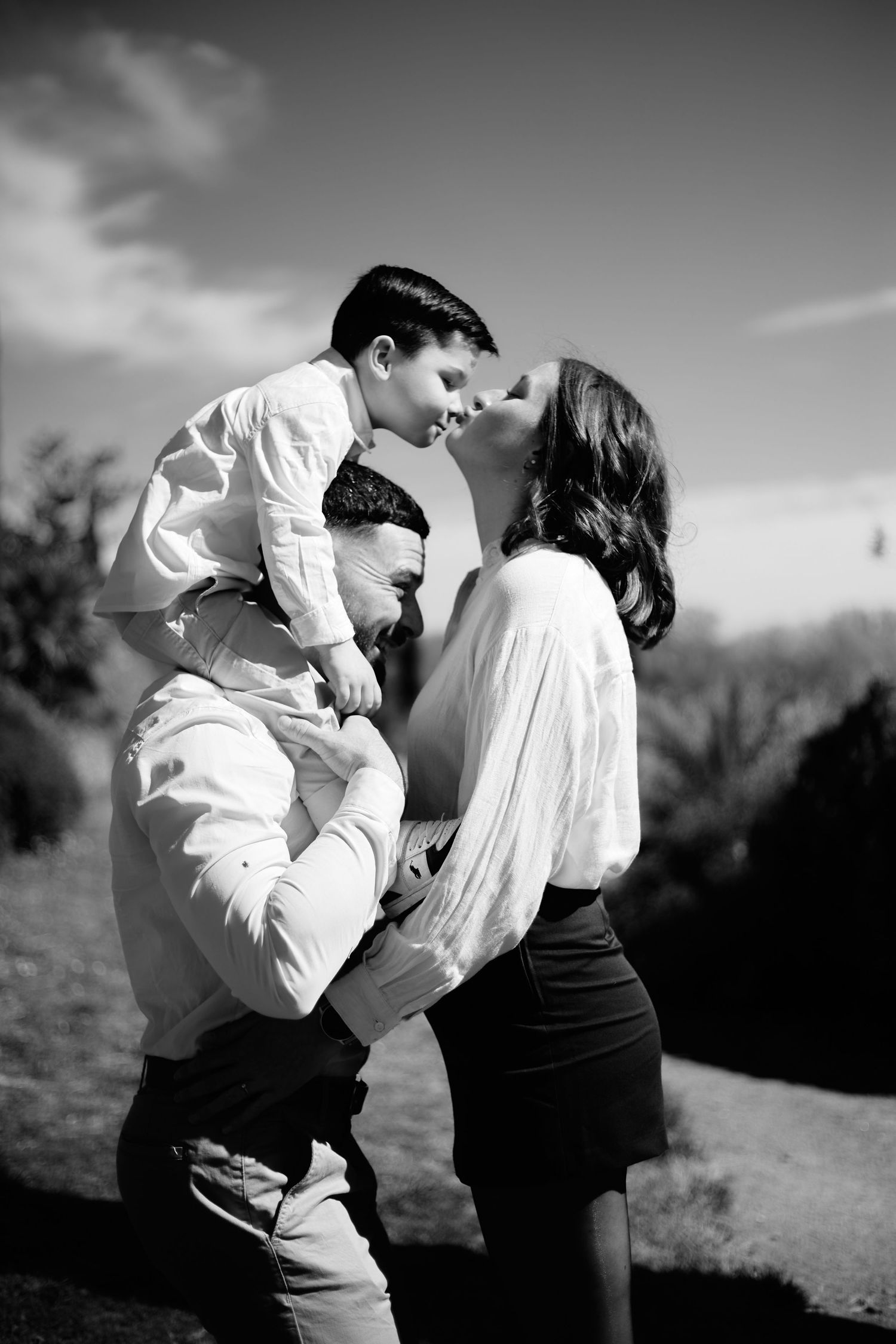 Black and white family photo of parents and young child on dad's shoulders sharing a tender moment outdoors.