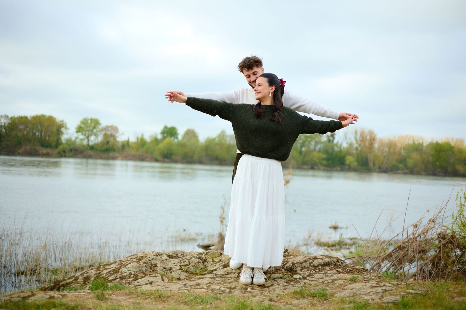Couple embracing by a lake with arms outstretched, surrounded by lush green trees under a cloudy sky.