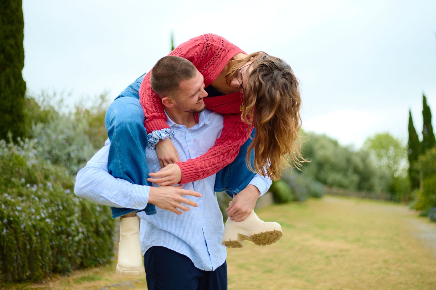Happy couple embracing outdoors, man lifting woman in red top, surrounded by lush green trees and garden path.