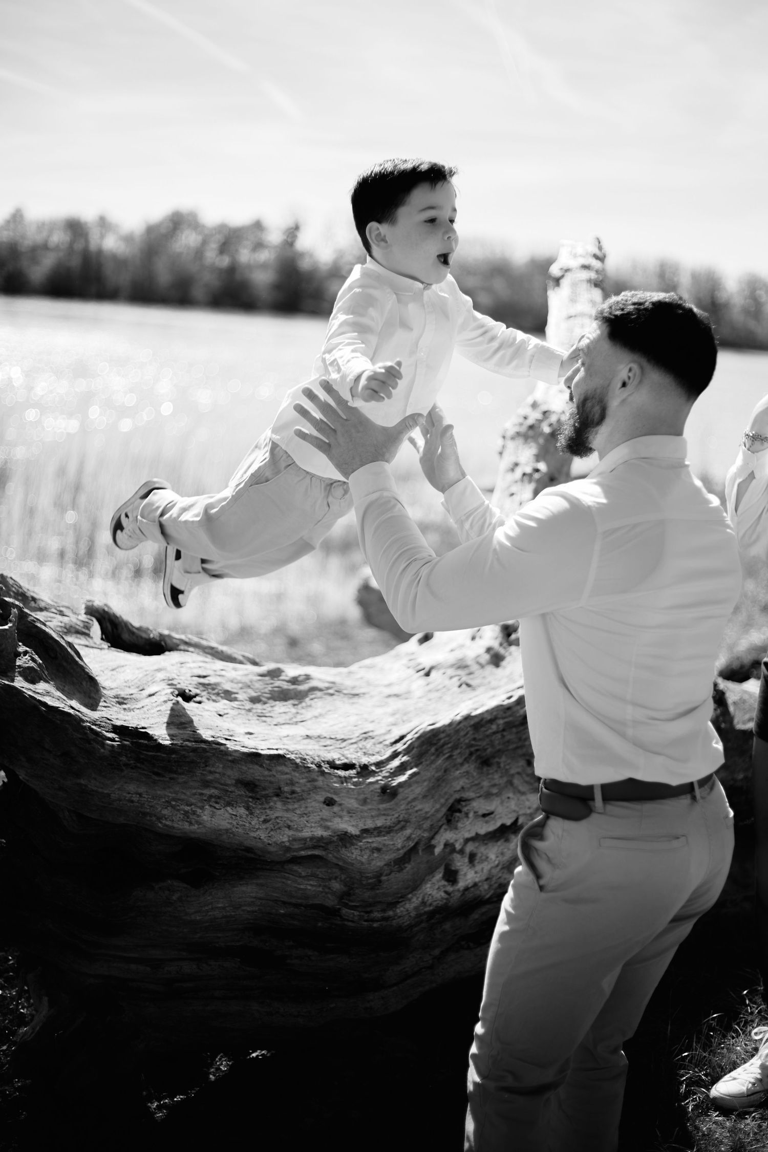 Father lifting young son in the air outdoors near a lake, both wearing white shirts, black and white photo.
