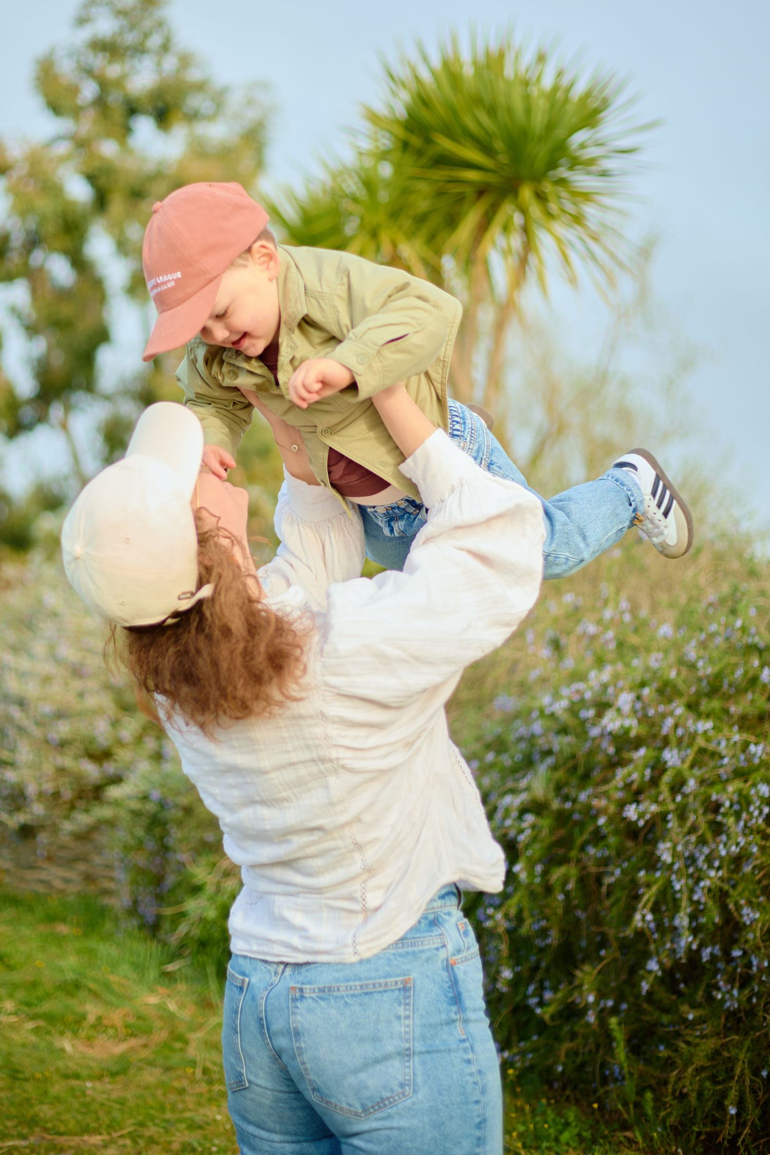 A mother lifts her toddler joyfully in a lush garden, both wearing caps and casual outfits on a sunny day.