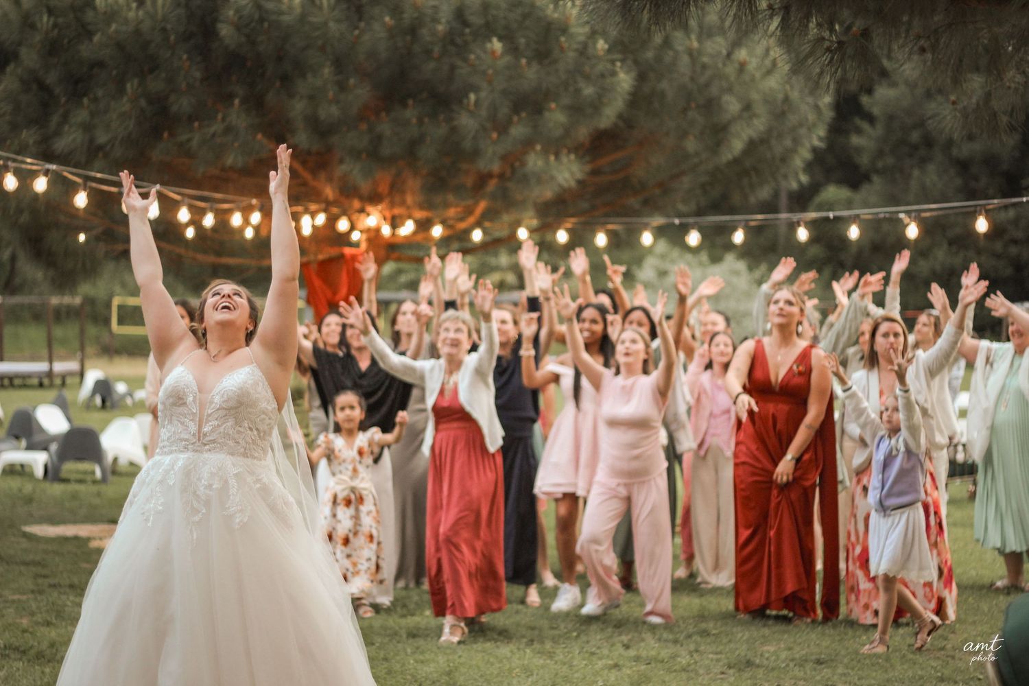 Large wedding party poses outdoors at sunset with string lights and coordinated dresses.