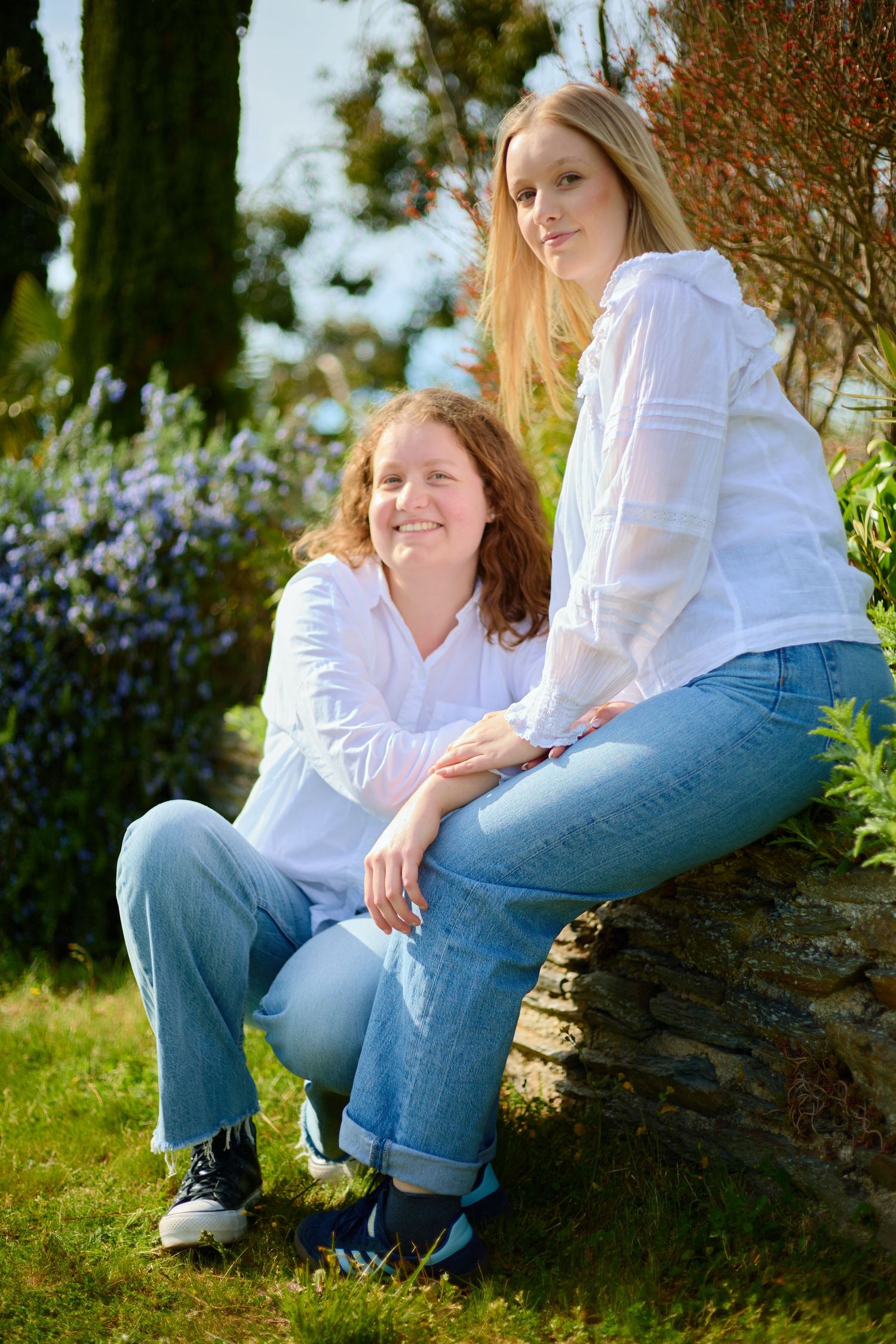 Two women in white blouses and jeans pose outdoors by a stone wall with purple flowers and trees.