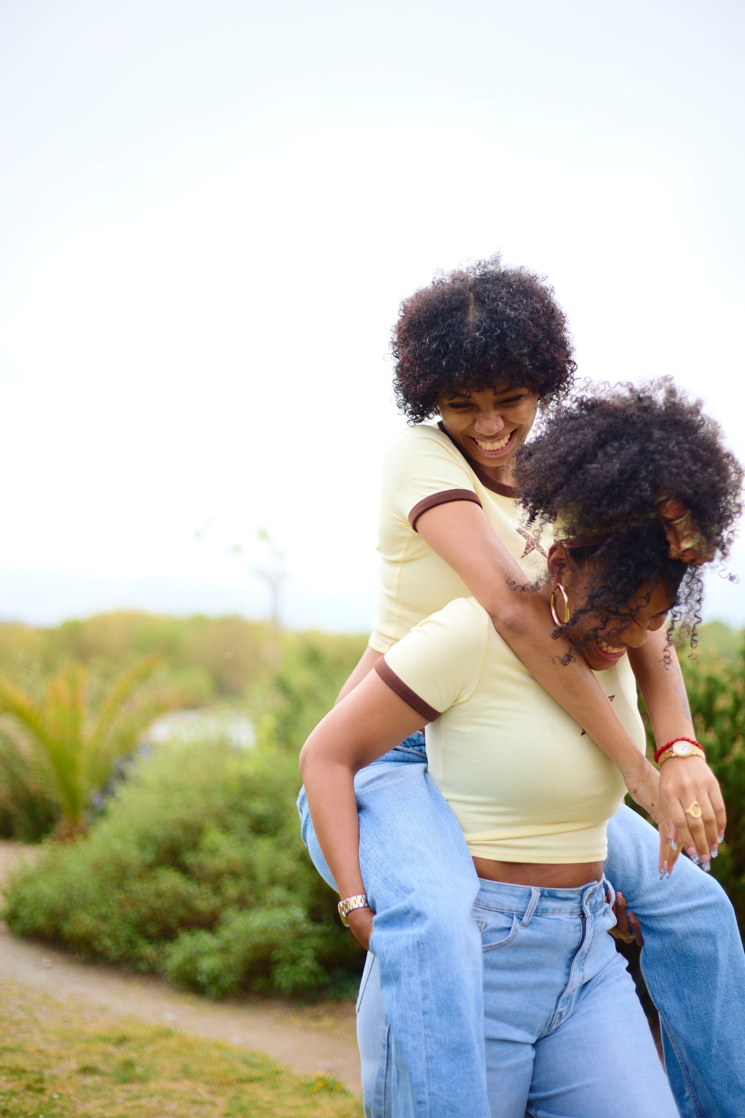 Two young Black women with afro hair laugh joyfully as one gives the other a piggyback ride outdoors.