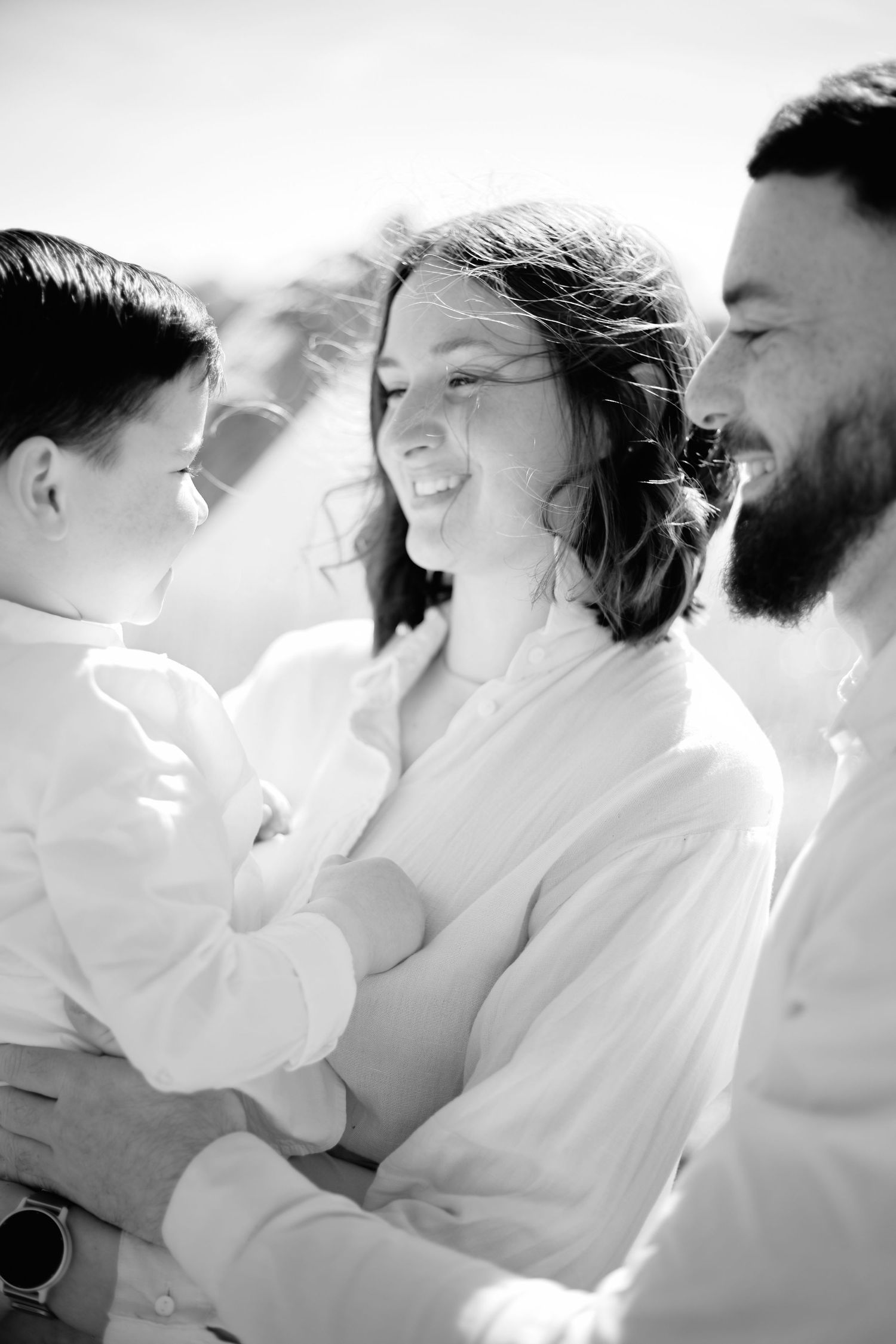 Black and white family portrait of smiling mother holding young son while bearded father looks on lovingly outdoors.