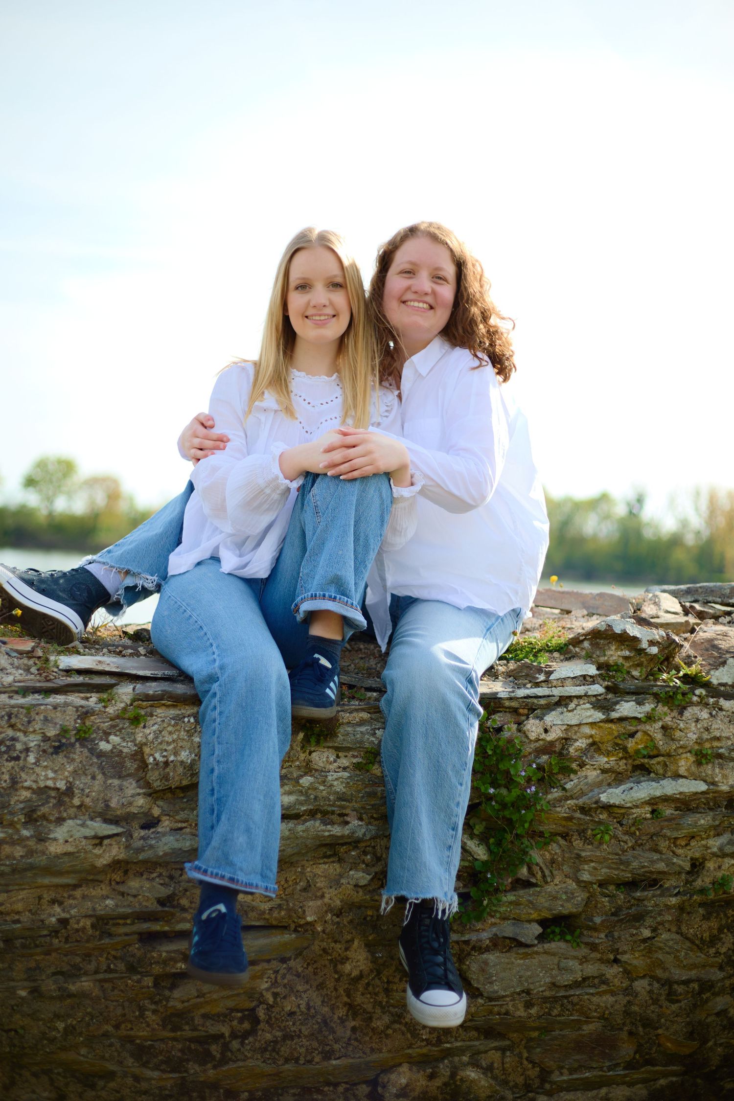 Two smiling women in white shirts and jeans sitting together on a stone wall outdoors on a sunny day.