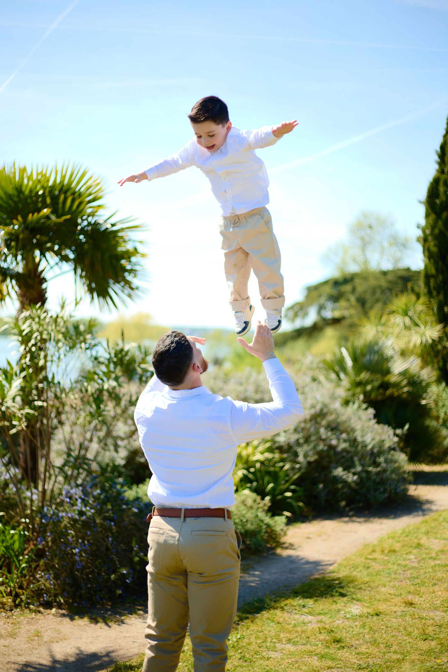 Father joyfully tosses young son into the air in a sunny garden with palm trees and blue sky.