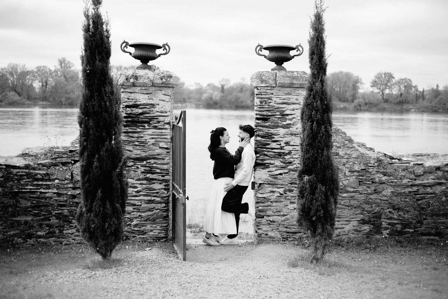 Black and white photo of a couple embracing in a stone gateway by a river, framed by cypress trees and decorative urns.