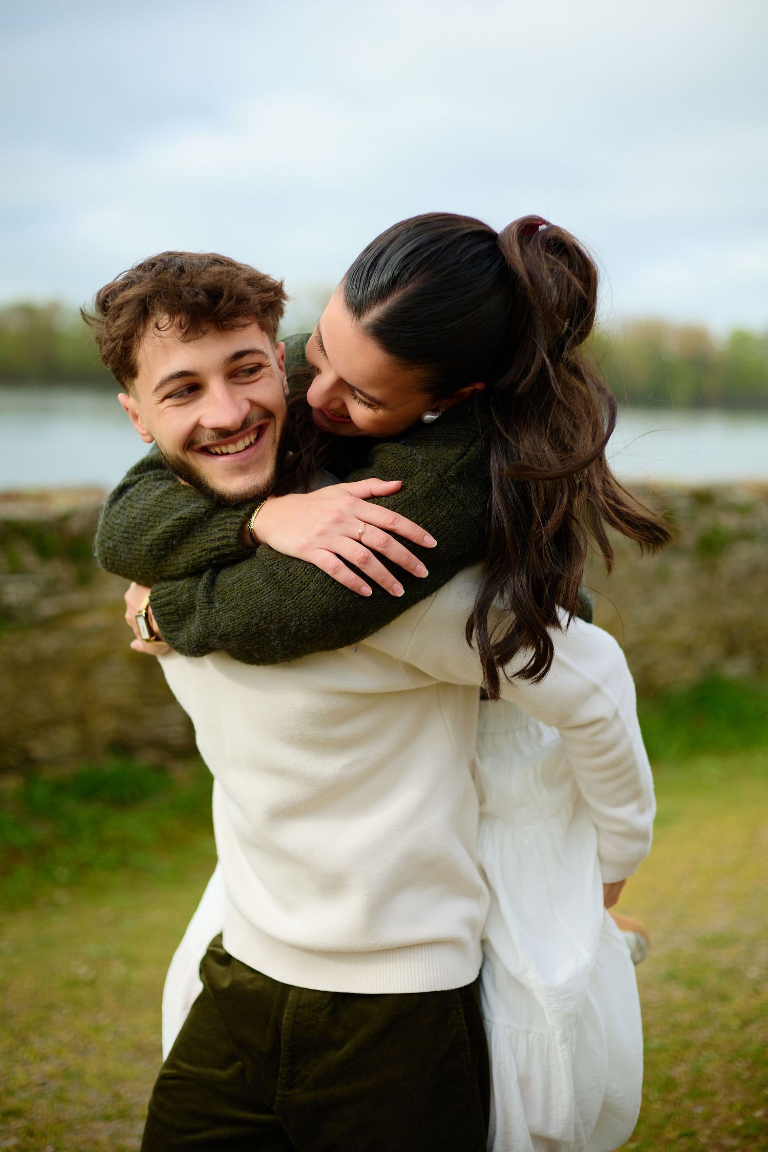 Happy couple embracing outdoors near a lake, smiling joyfully in casual green and white sweaters.