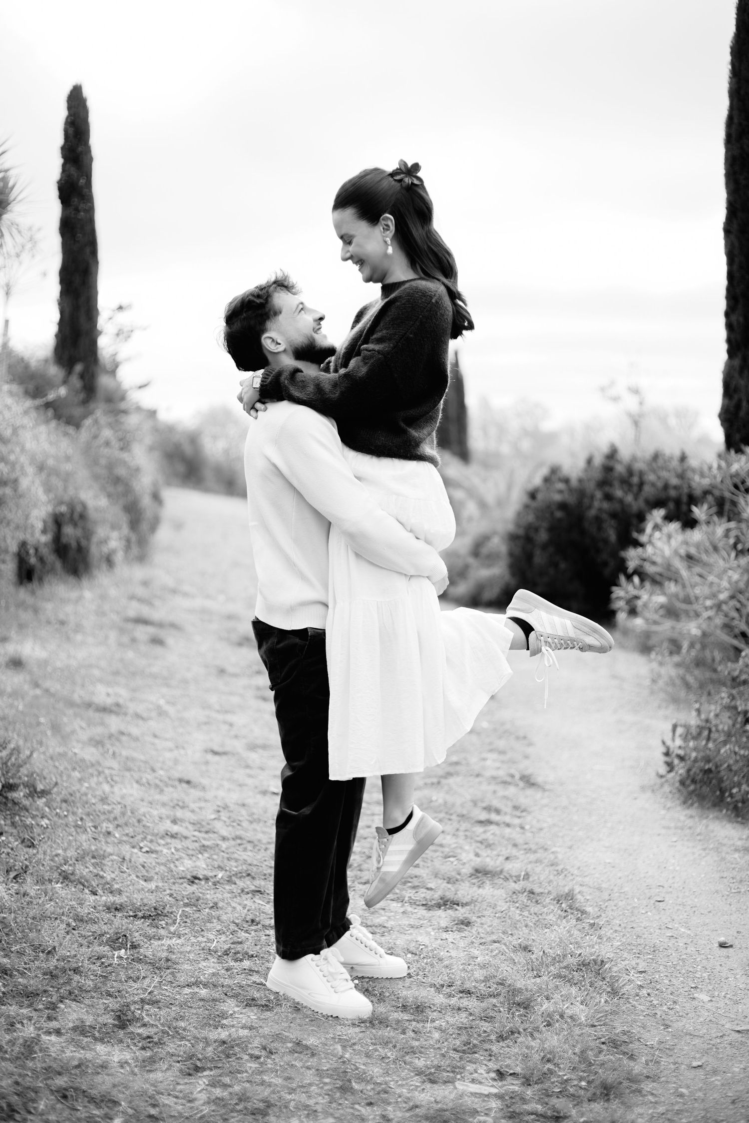 Black and white photo of a man lifting a smiling woman outdoors on a garden path, sharing a romantic moment.