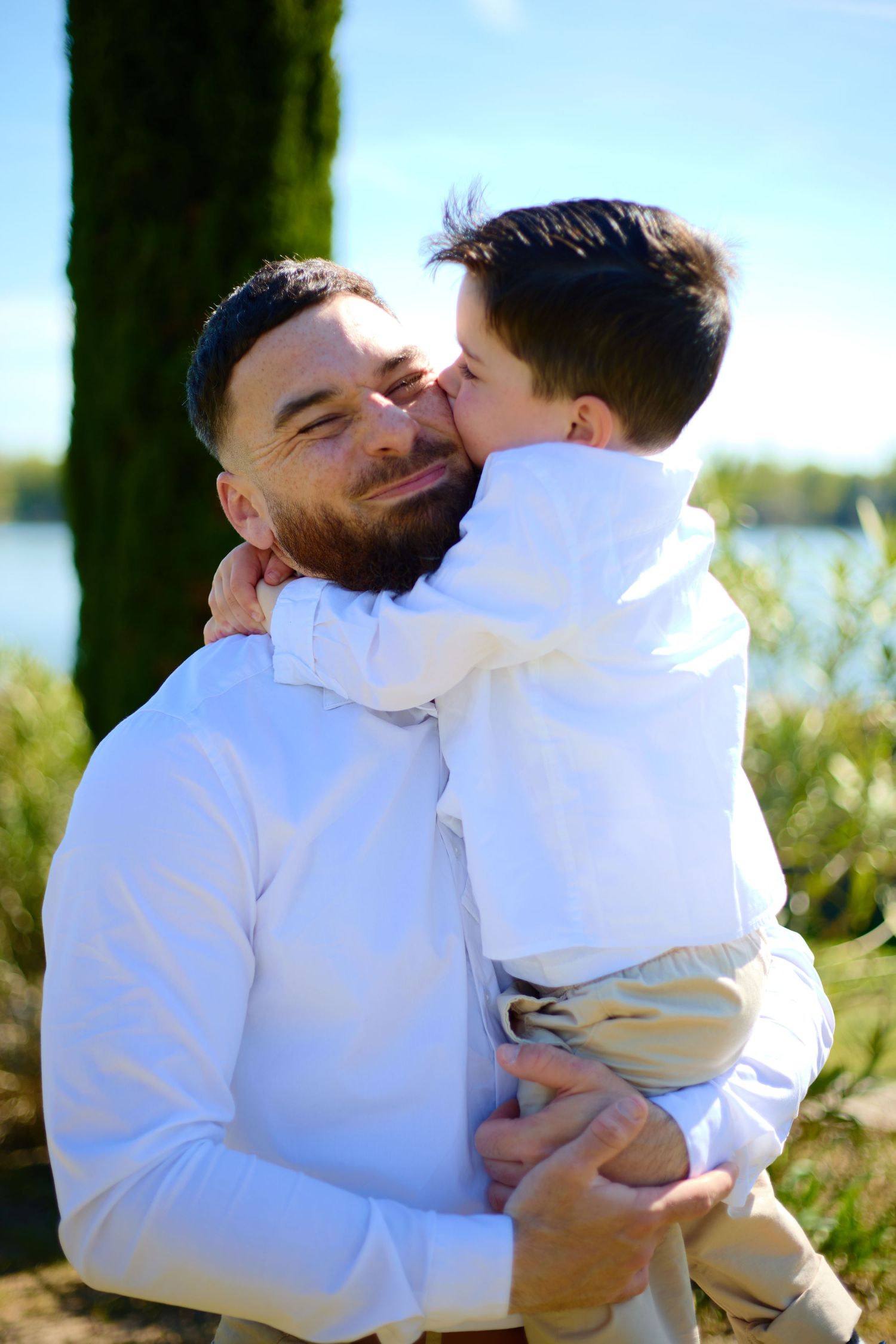 A bearded father in white shirt holds his young son who kisses his cheek outdoors near a lake.