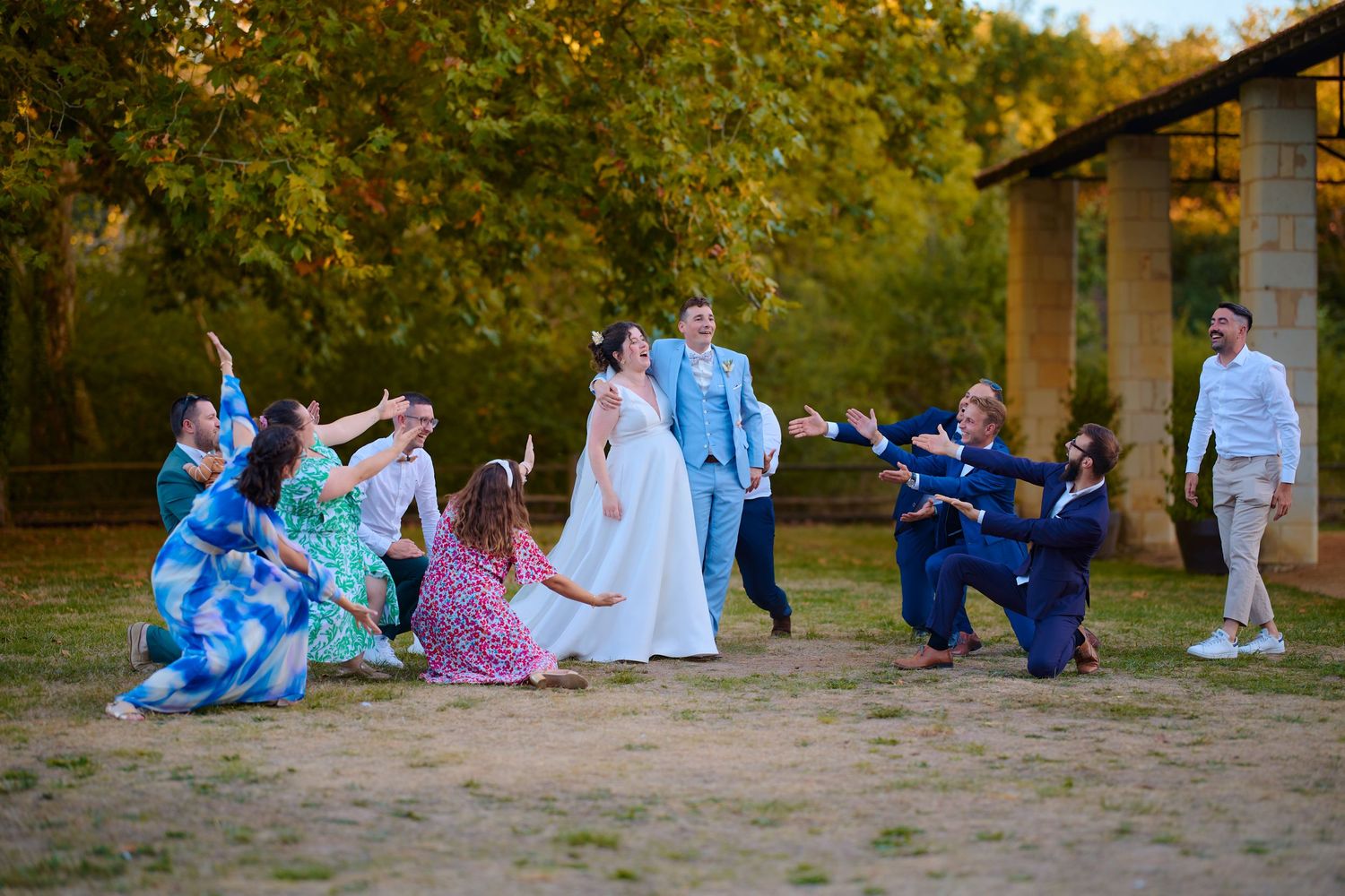 Wedding couple surrounded by joyful guests striking fun poses outdoors near trees at golden hour.