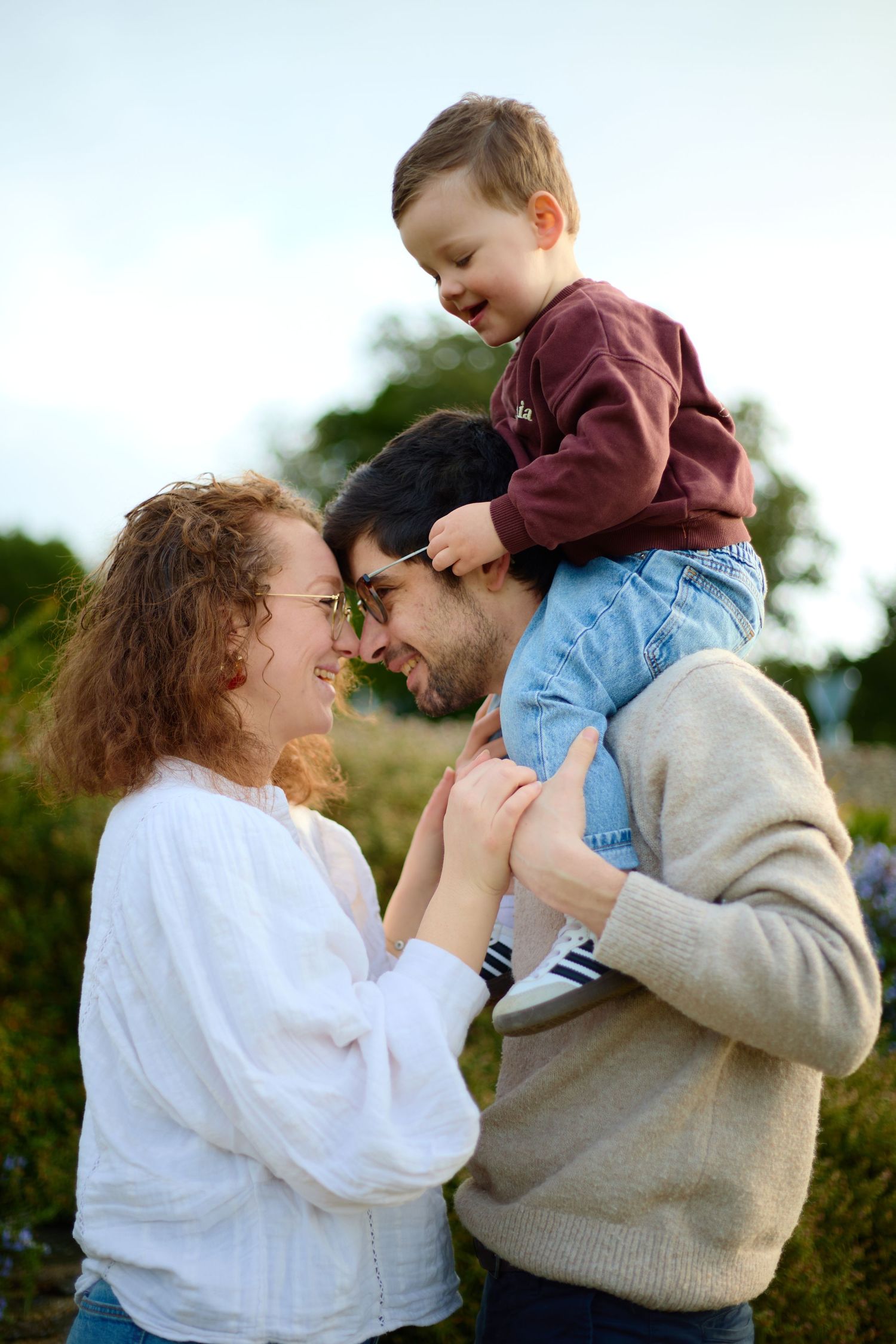 A happy young family outdoors, mother with curly red hair smiling at father carrying toddler on shoulders.