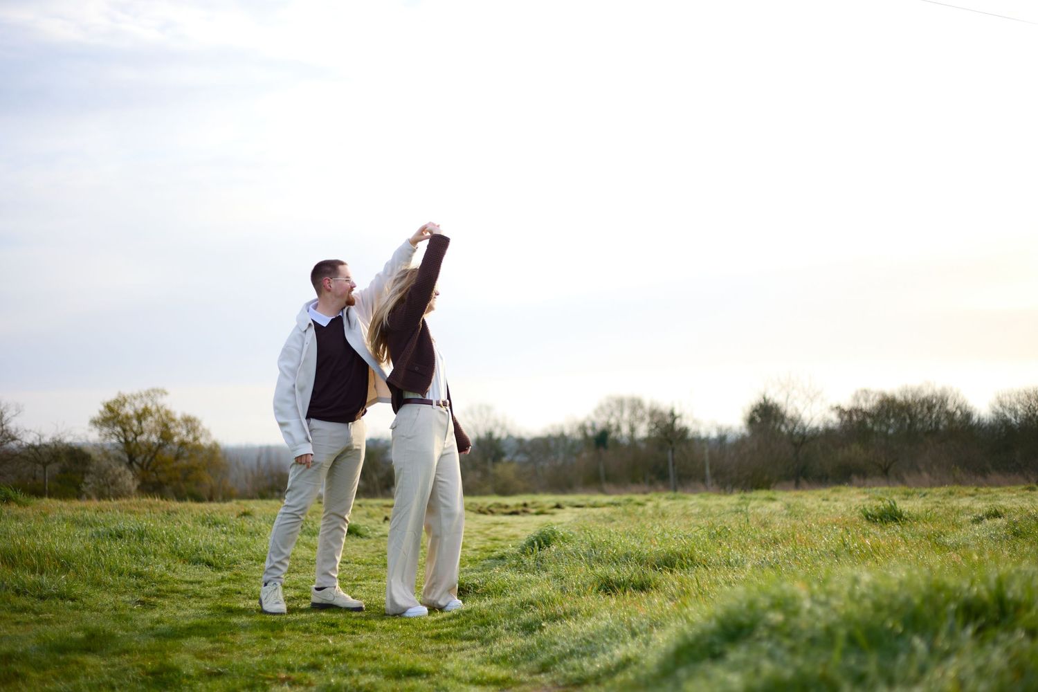 Couple strolling hand in hand across a sunlit grassy field with trees in the background.