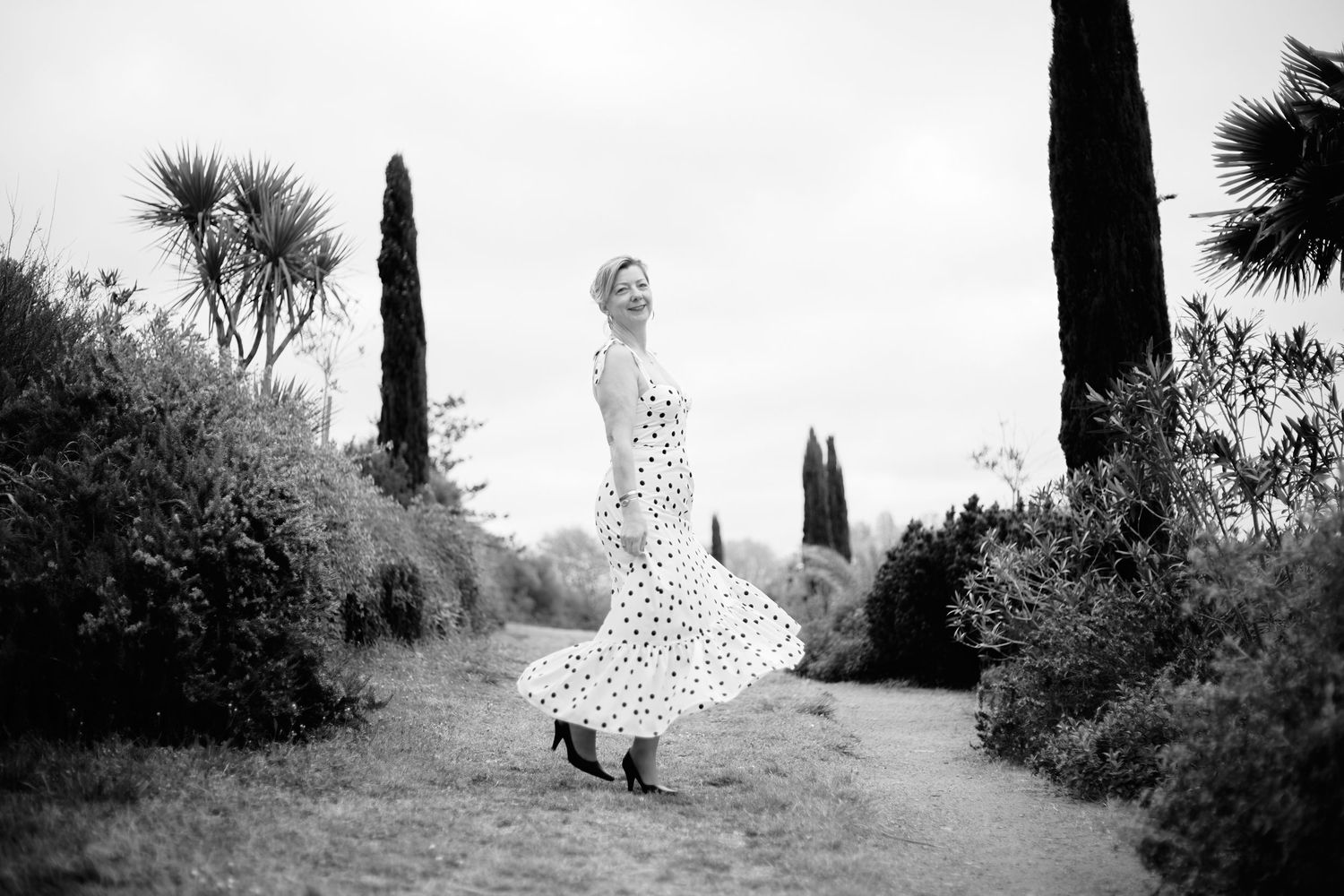 Woman in polka dot dress twirling on garden path with palm and cypress trees in black and white photo.