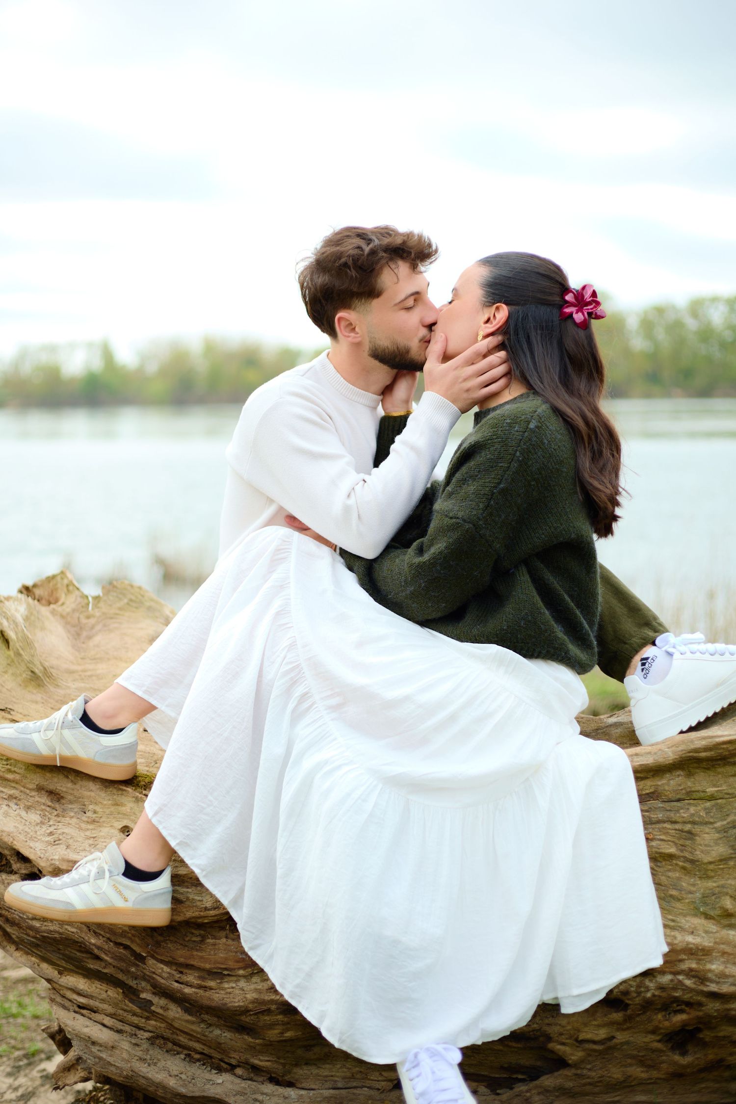 Couple sharing a romantic kiss while sitting on a large log by a serene lake, woman in green sweater and white skirt.