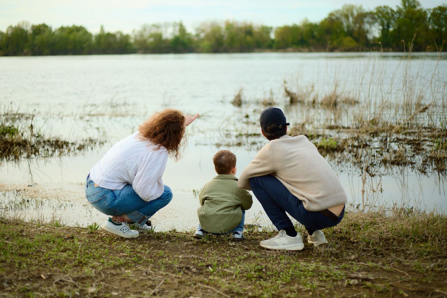 Family crouching by a lake shore, pointing at water, surrounded by lush greenery on a calm day.