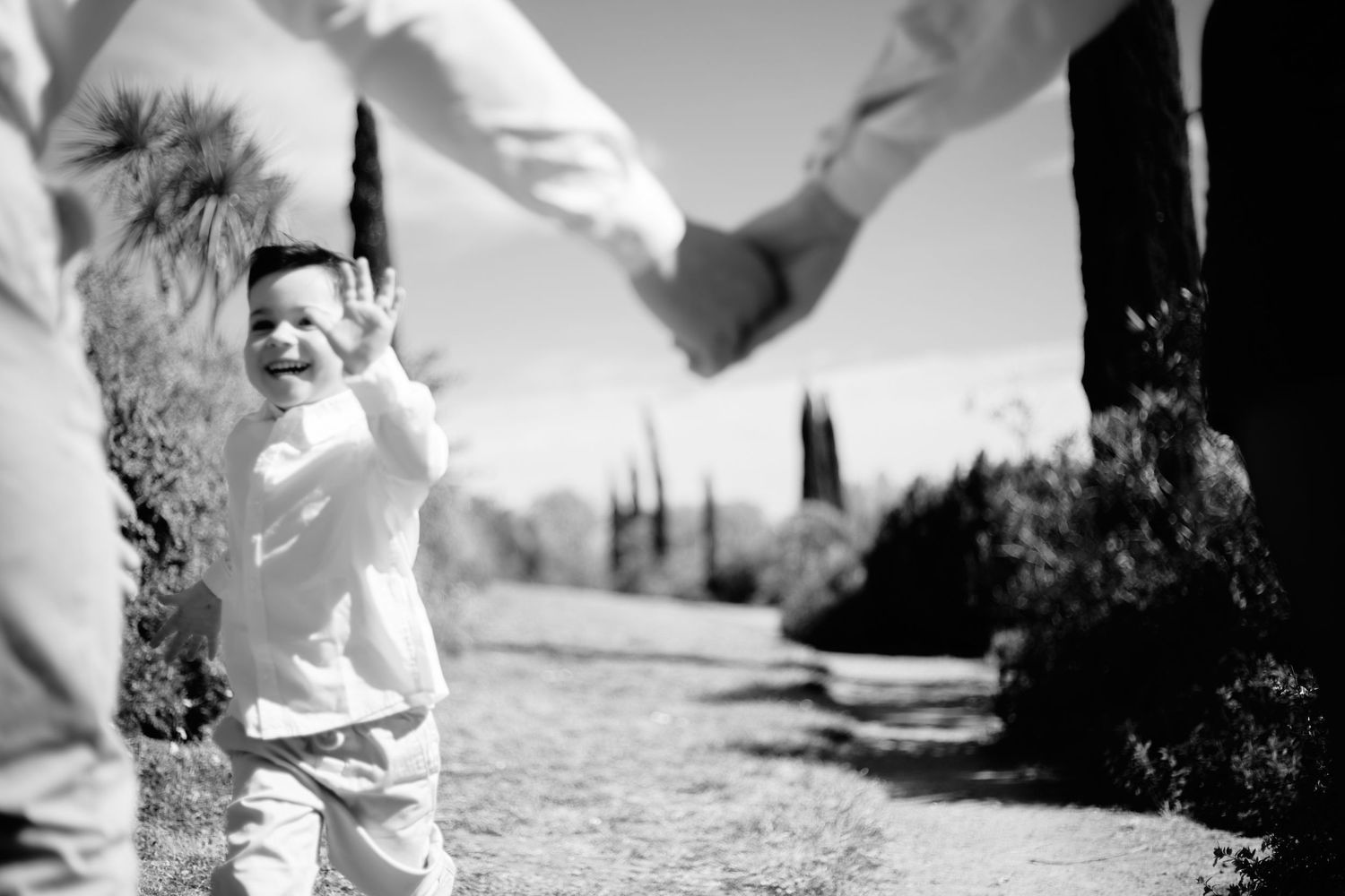 Black and white photo of a toddler walking between two adults holding hands on a garden path.