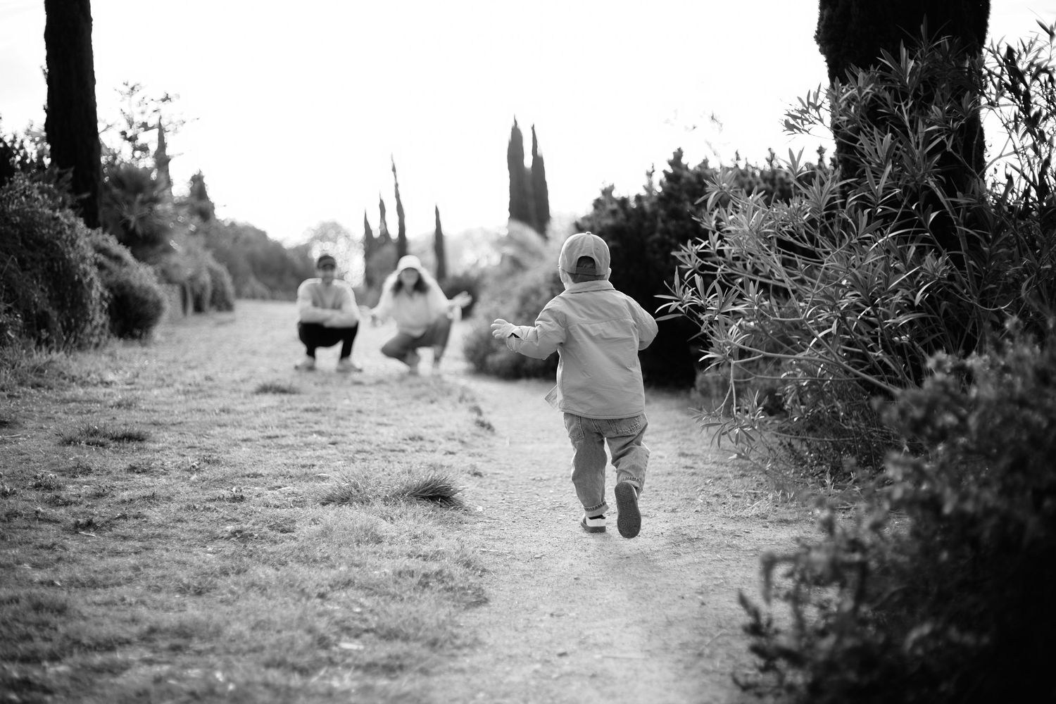 Black and white photo of a toddler running down a path toward a crouching adult in a lush outdoor setting.